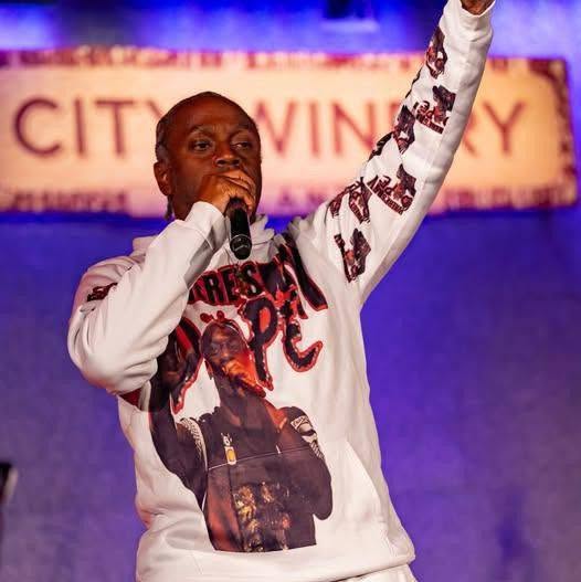 Person speaking into microphone with arm raised, wearing a sweatshirt with a large graphic and text, on stage with a blurred background.