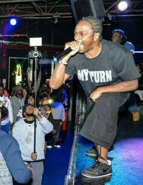 Performer with gray hair, black T-shirt, and shorts, holding microphone on stage with audience in background.