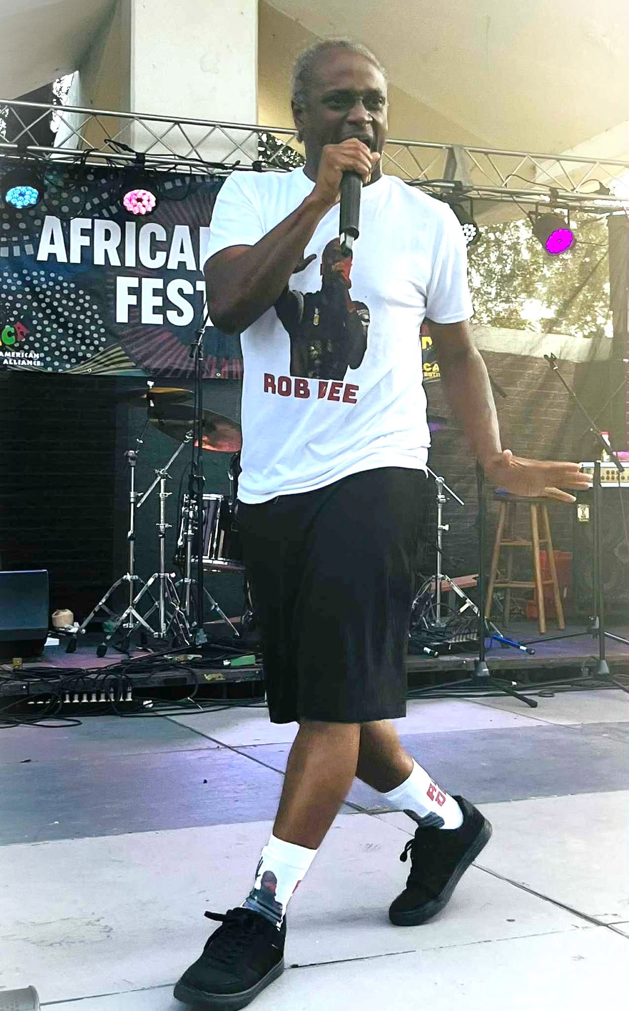 Person holding microphone on stage with African Festival banner in background, wearing white T-shirt, black shorts, and sneakers.