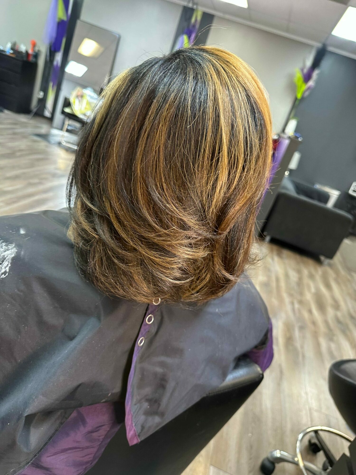 Top view of a person's head with styled, layered hair in shades of brown and blonde, sitting in a salon chair.