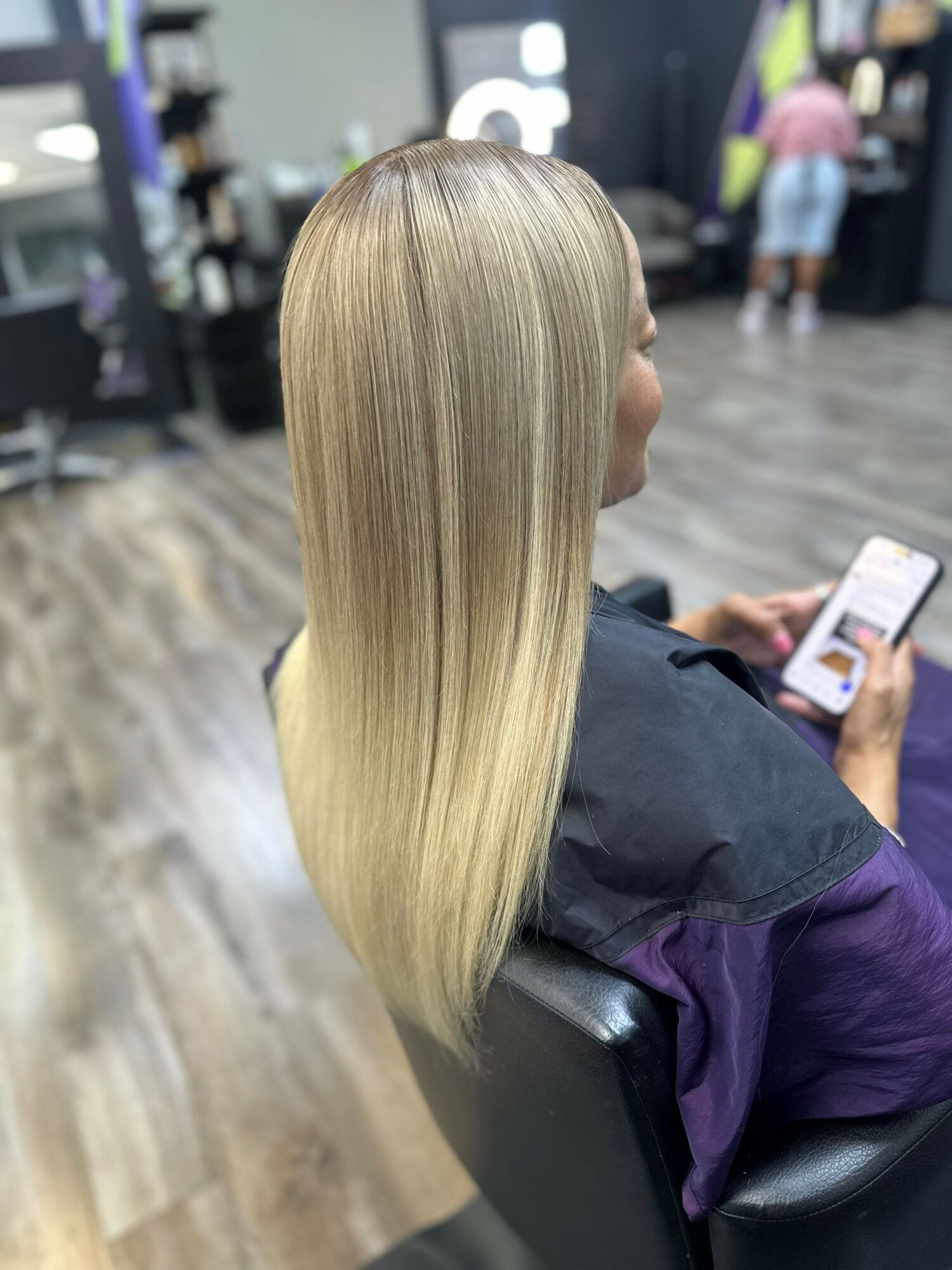 Person with long, straight, blonde hair sitting in a salon chair, looking at a phone, with salon equipment in background.