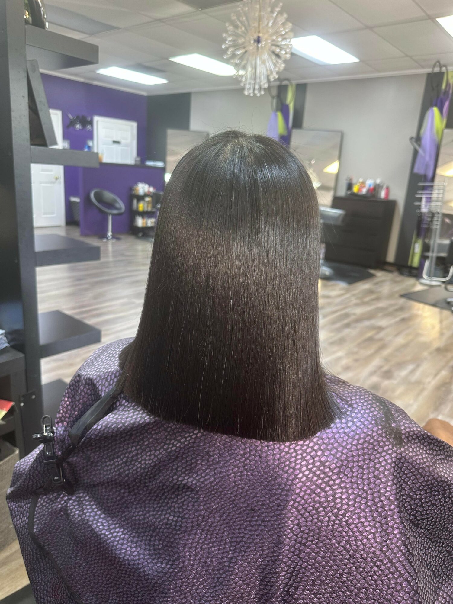 Person with straight, shiny, shoulder-length hair sitting in a salon chair, facing away, with salon interior in background.