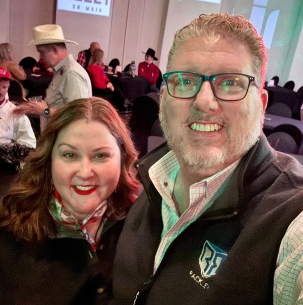 Two people smiling at an indoor event, with a woman on the left and a man on the right wearing glasses and a jacket.