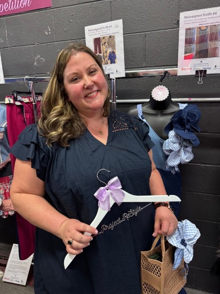 Woman with shoulder-length hair smiling, holding a ribbon, standing in front of clothing displays, with a basket nearby.