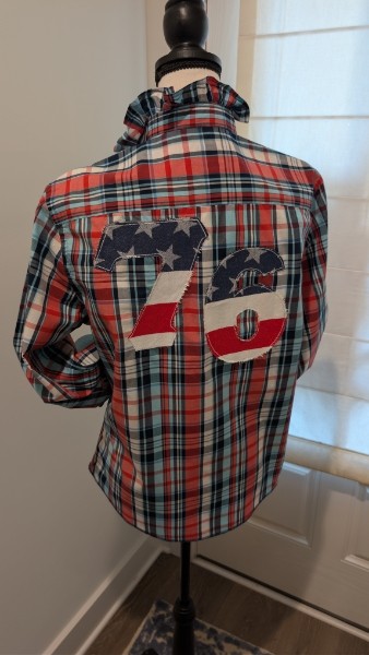 Back of a plaid shirt with patches, displayed on a mannequin, near a window with curtains.