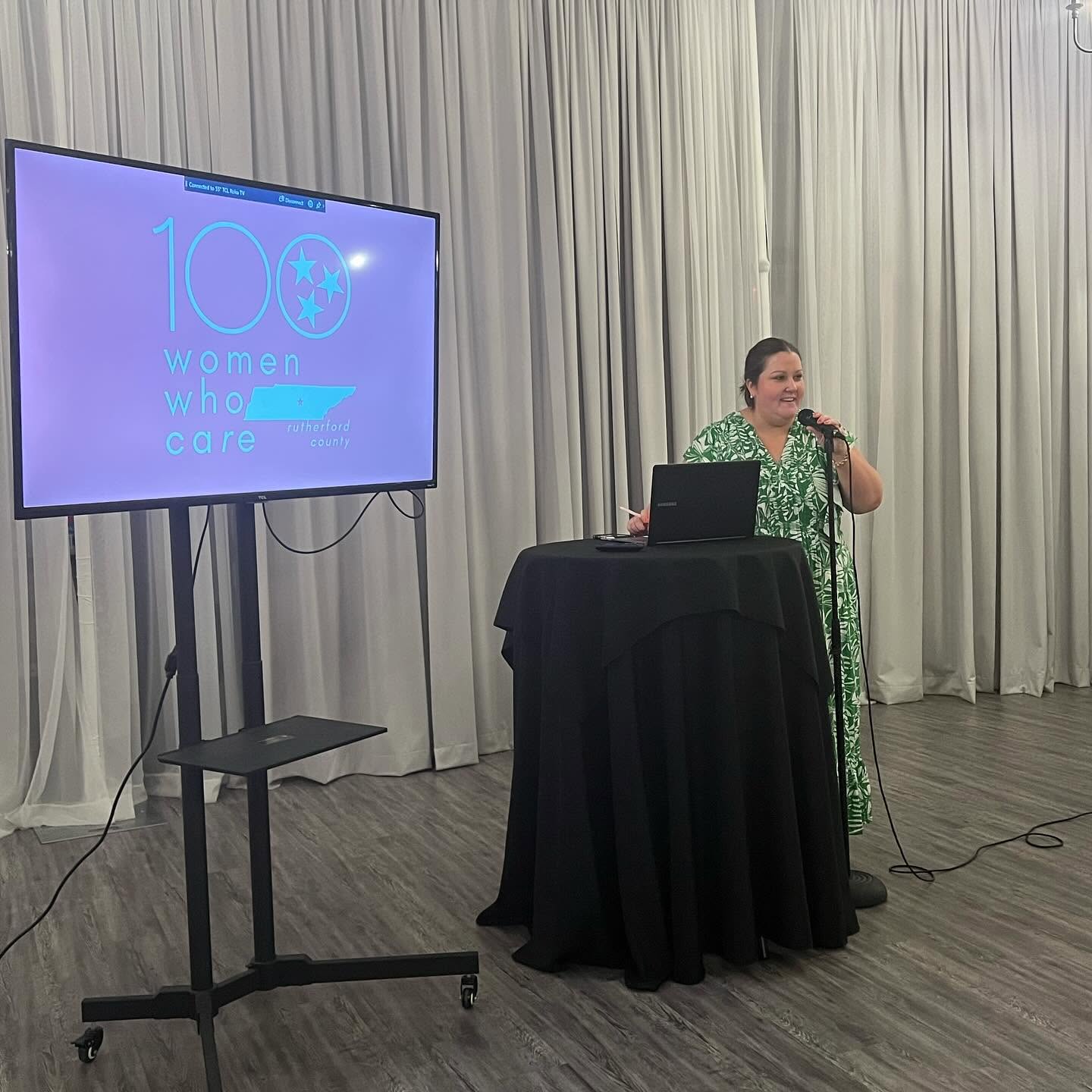 Woman speaking into microphone at a presentation event, standing next to a table with a laptop, in front of a large screen displaying a logo and text.