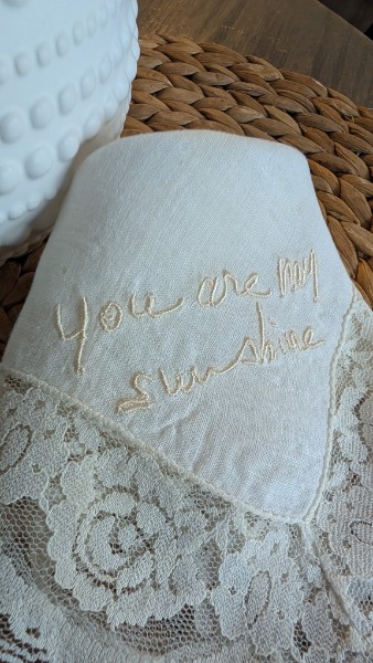 A white cloth with gold embroidered text reading 'You are my sunshine' on lace fabric, placed on a woven surface.