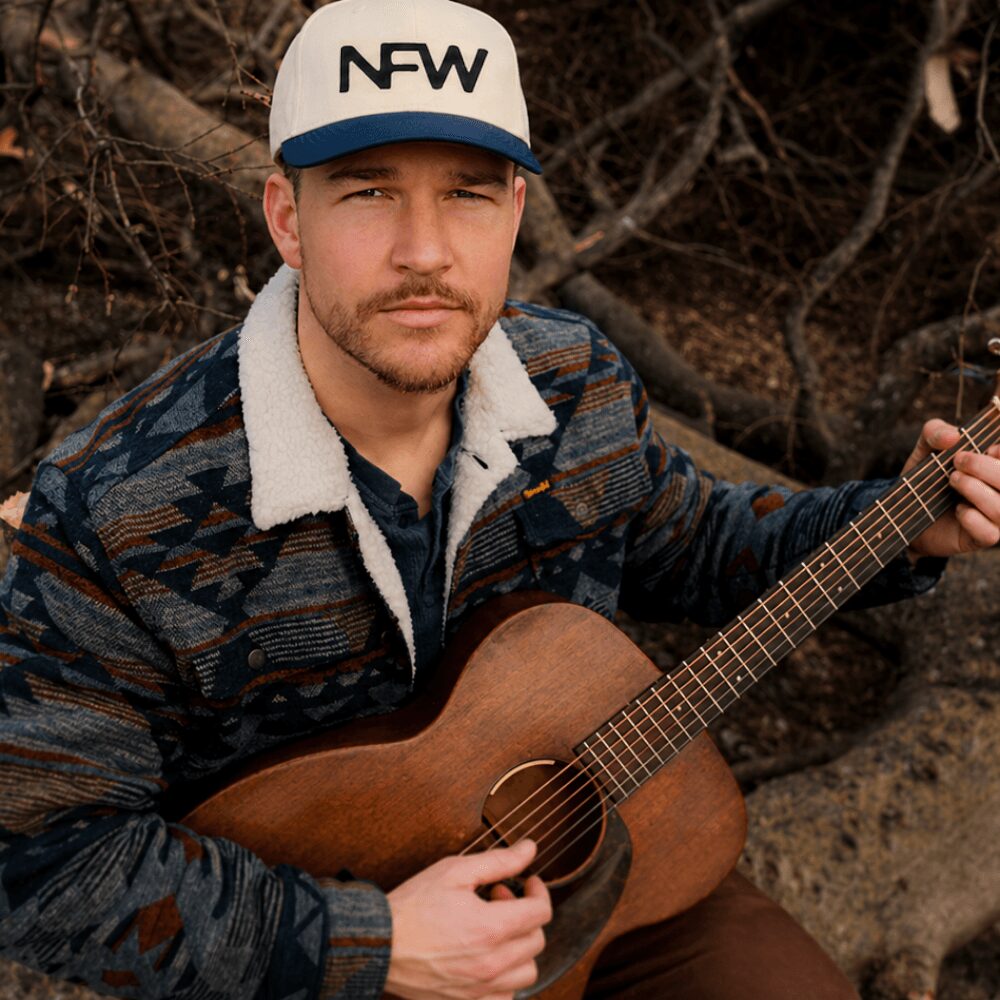 Man wearing a white cap with 'NFW' logo, holding a guitar outdoors, with a background of trees and rocks.