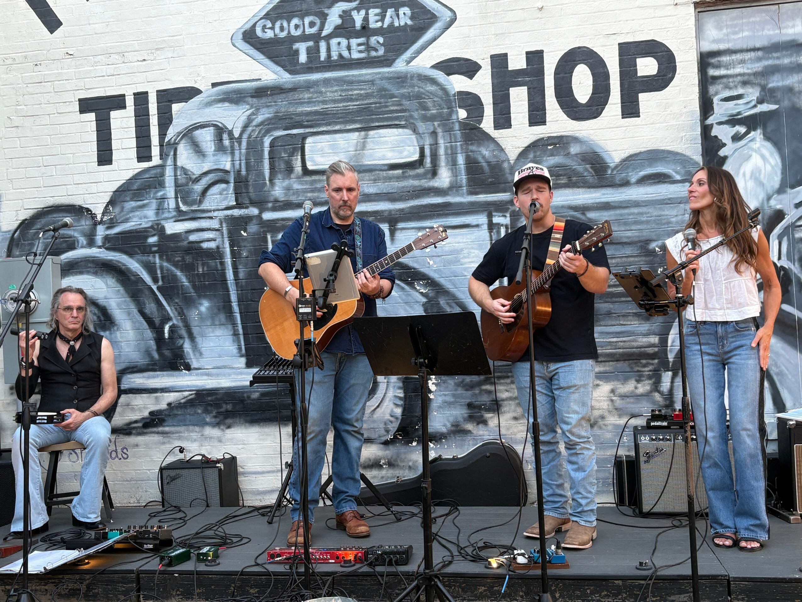 Three musicians perform on stage with guitars, a woman sings, and a man plays keyboard, in front of a mural of vintage trucks.