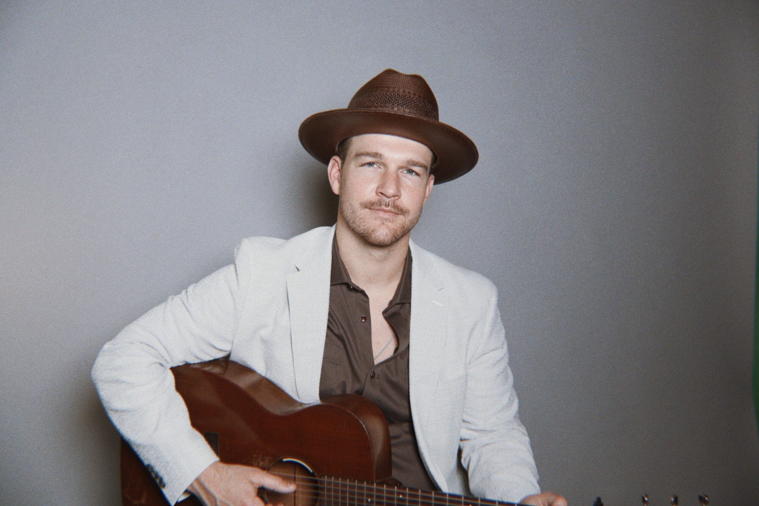 Young man with a hat holding a guitar, wearing a white jacket and brown shirt, standing against a gray background.