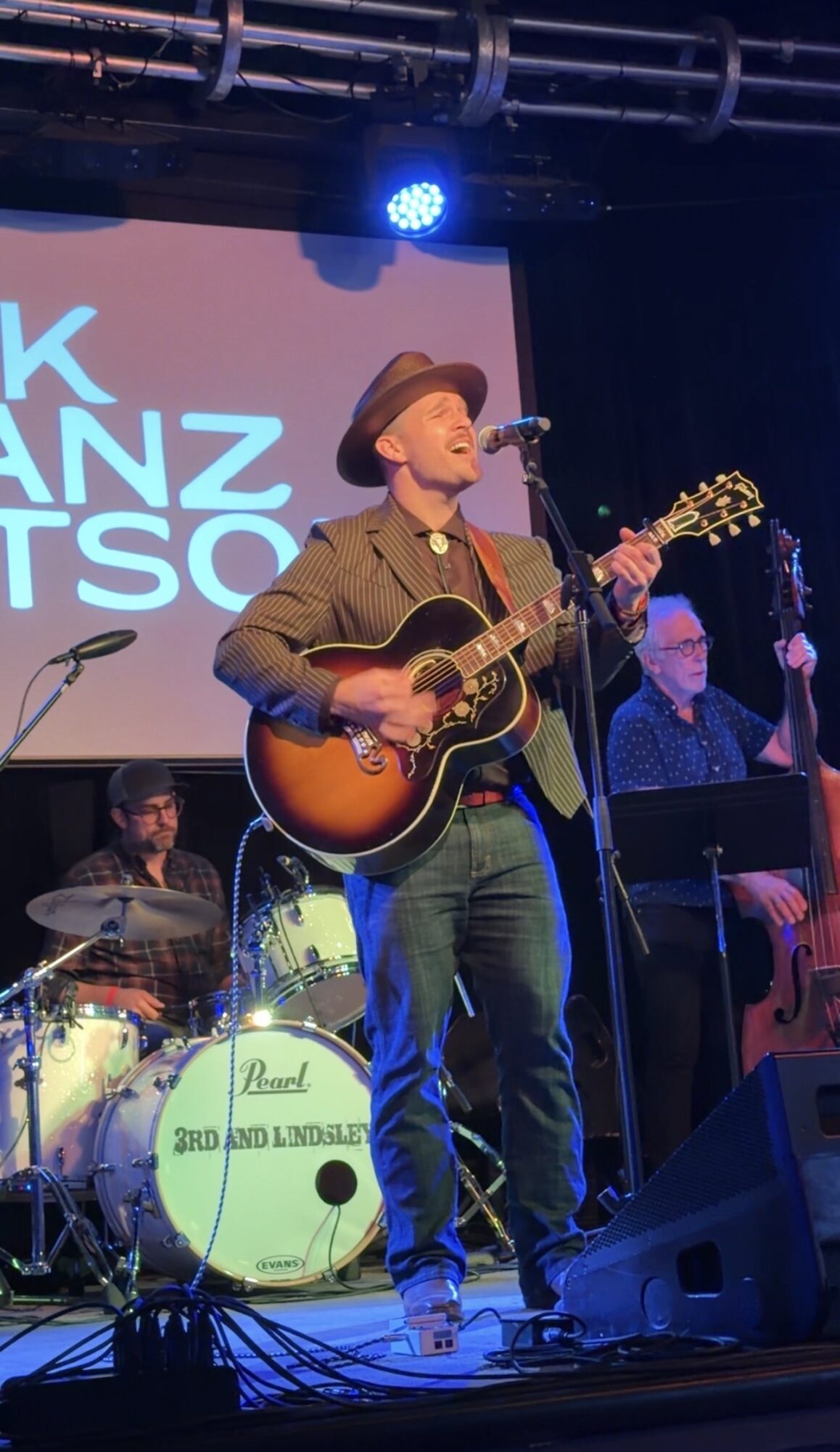 Musician with hat playing guitar on stage, band members and drum set visible, stage lighting overhead.