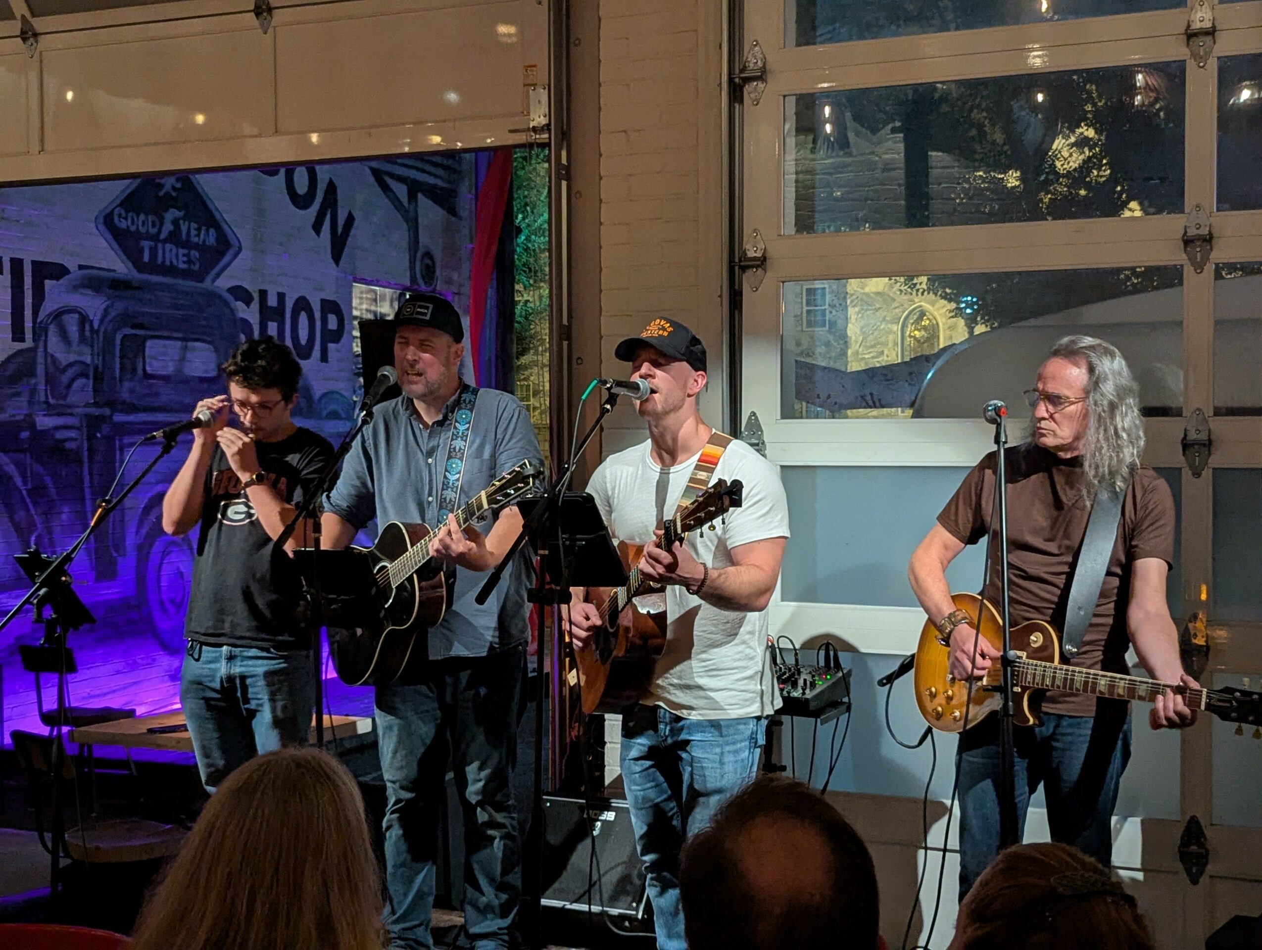 Four musicians perform on stage with guitars, microphones, and a colorful background, audience visible in foreground.