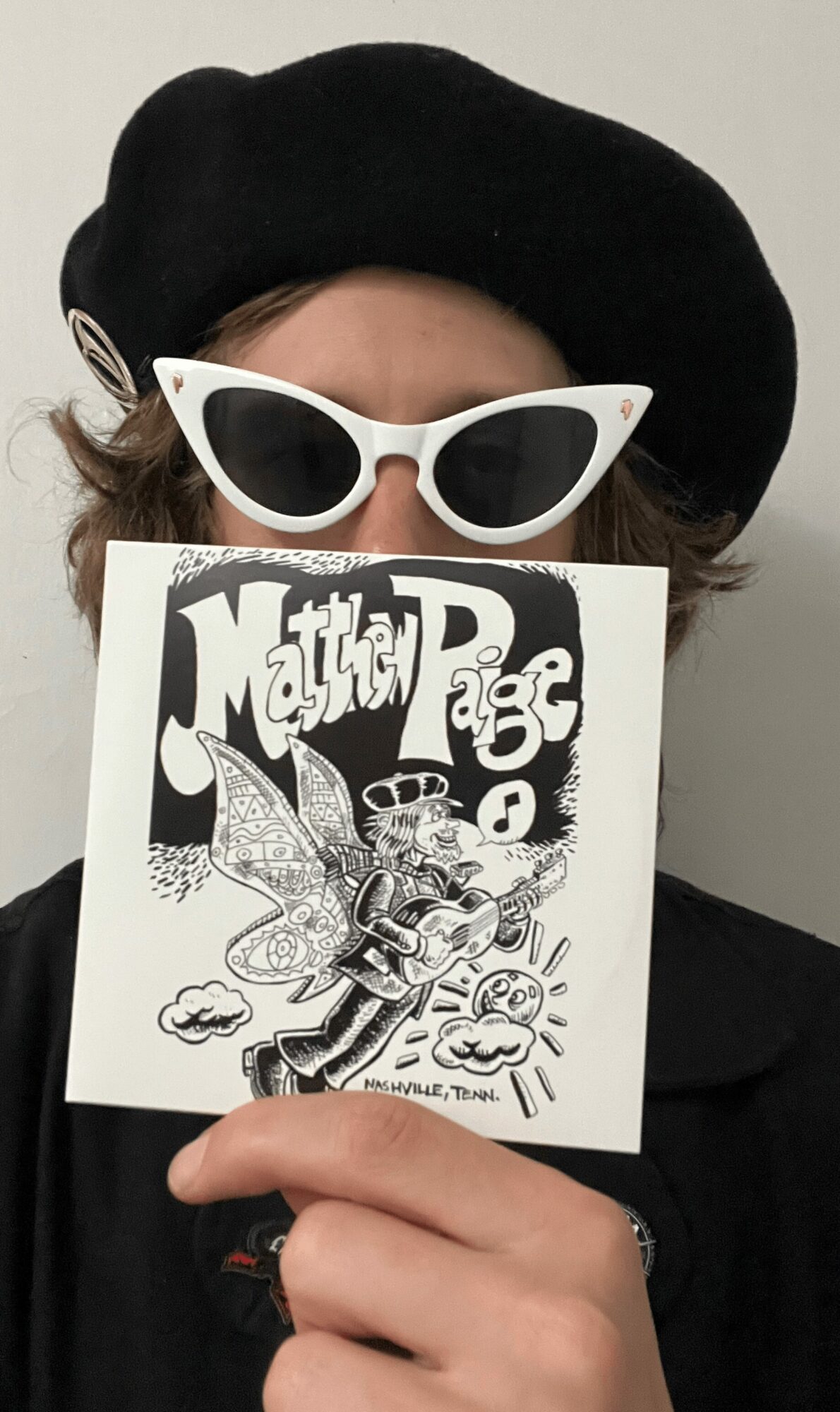 Person wearing a black beret and white sunglasses, holding a black-and-white drawing in front of their face.