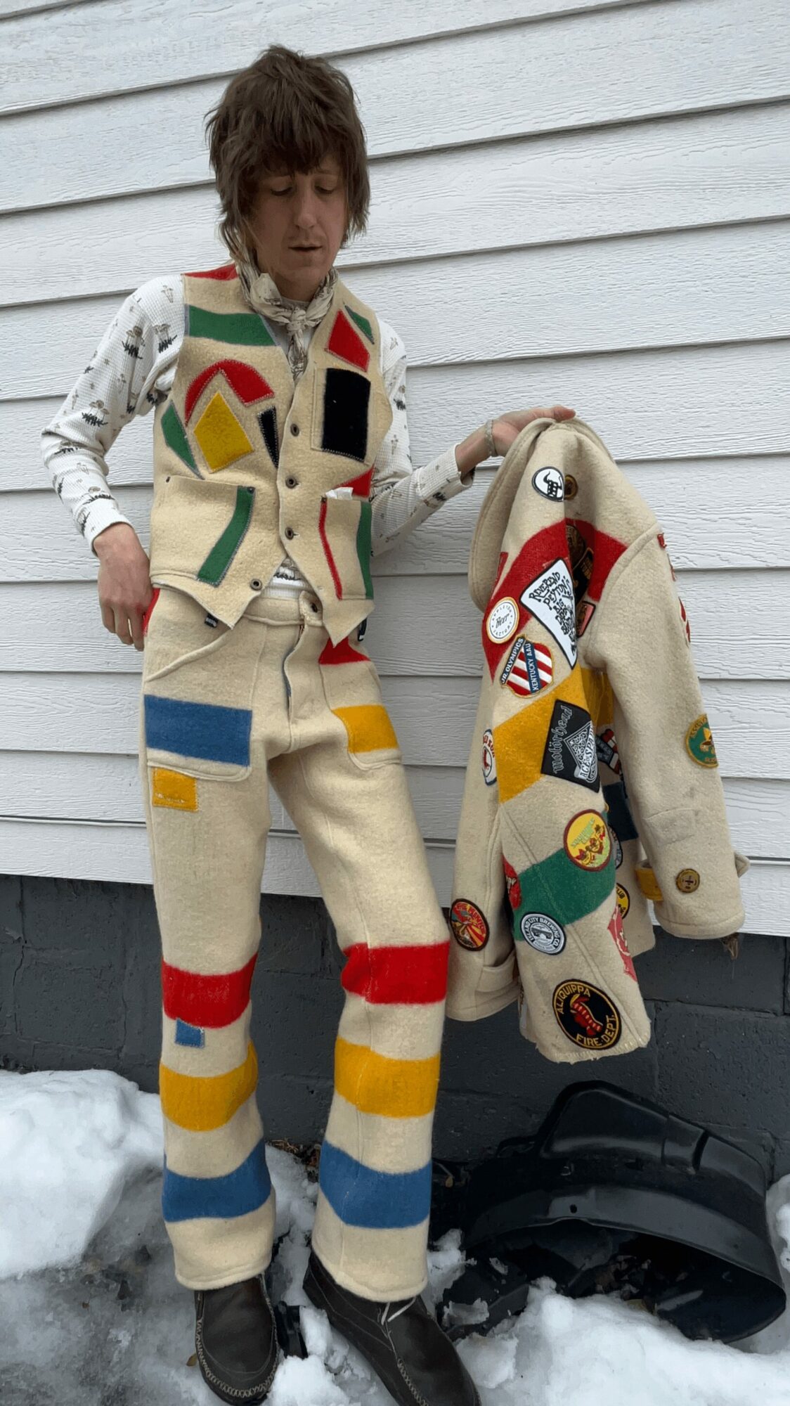 Young person standing outdoors in snow, wearing colorful patchwork pants and holding a matching patchwork coat, with a white house in background.
