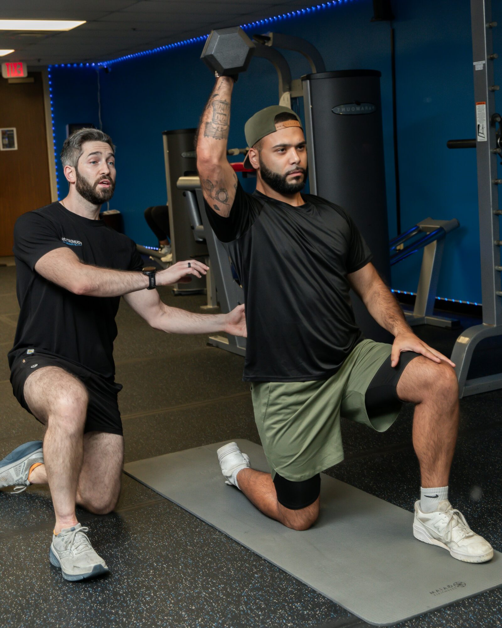 Personal trainer assists man performing a lunge with a dumbbell in a gym. Both are focused on exercise. Word count: 20.