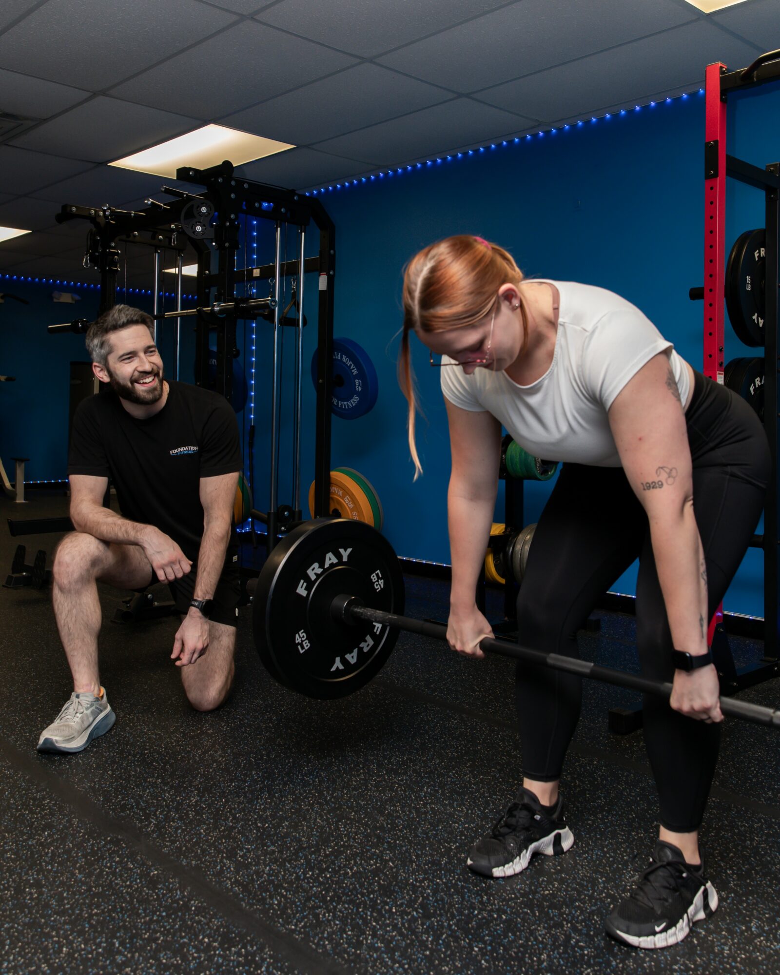 Woman lifting a barbell with a man watching and smiling in a gym setting.
