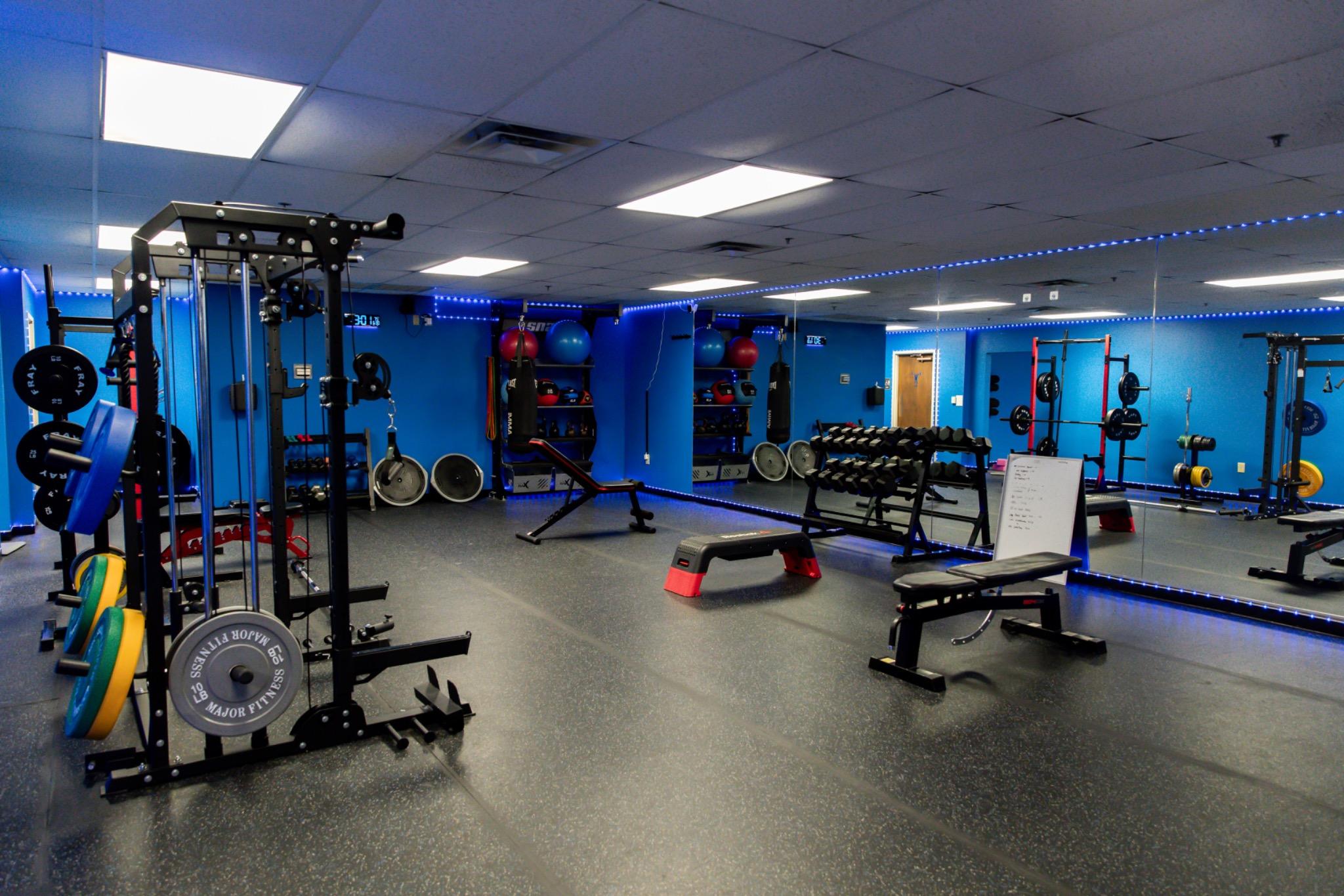 Empty gym with weightlifting equipment, benches, and mirrors, illuminated by ceiling lights and blue wall accents.