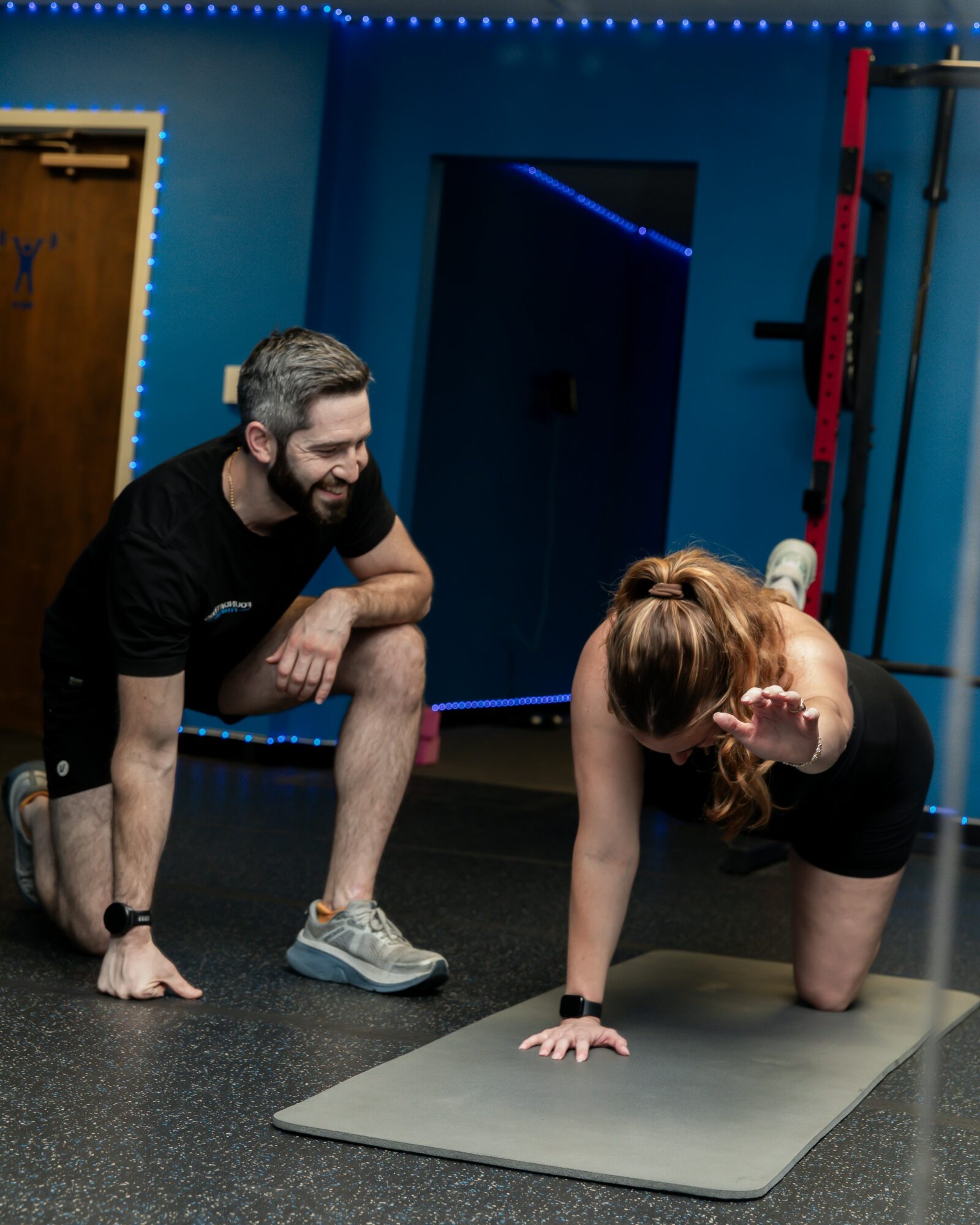 A man and woman exercising on a mat in a gym, with the woman on her hands and knees, the man smiling nearby.