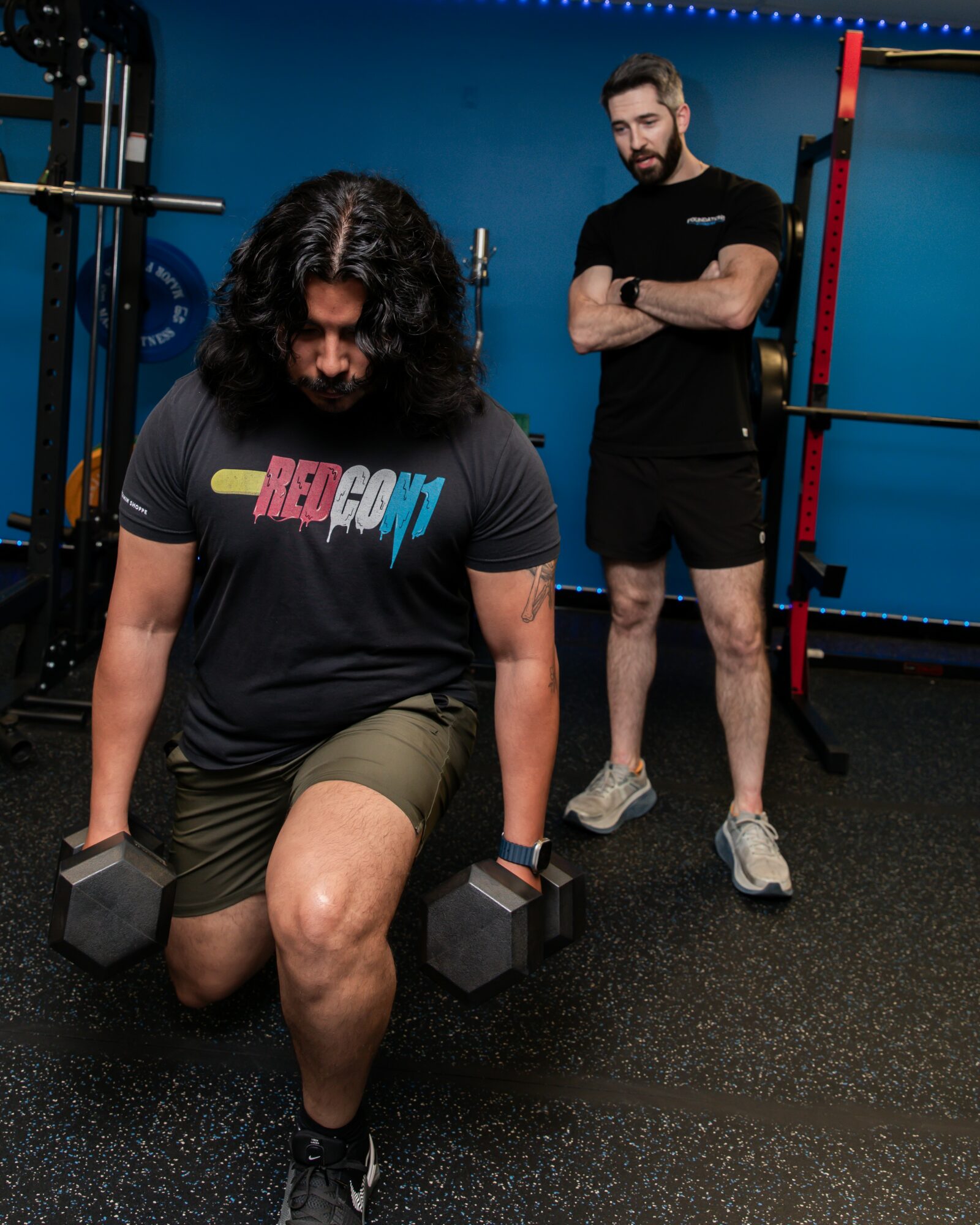 Person kneeling with dumbbells in a gym, another person standing with arms crossed behind, gym equipment visible.