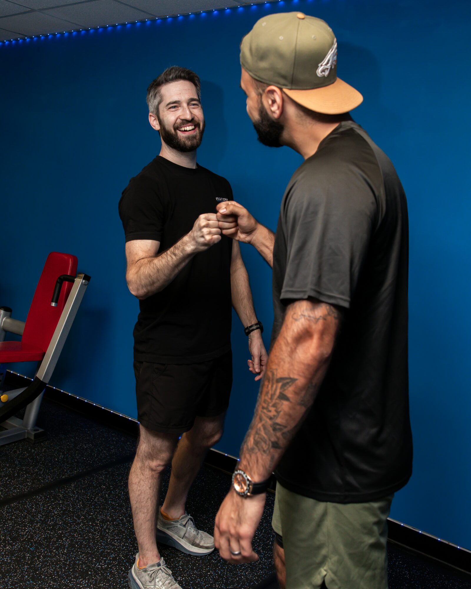 Two men smiling and shaking hands in a gym with a blue wall, one wearing a black shirt and shorts, the other in a cap and t-shirt.