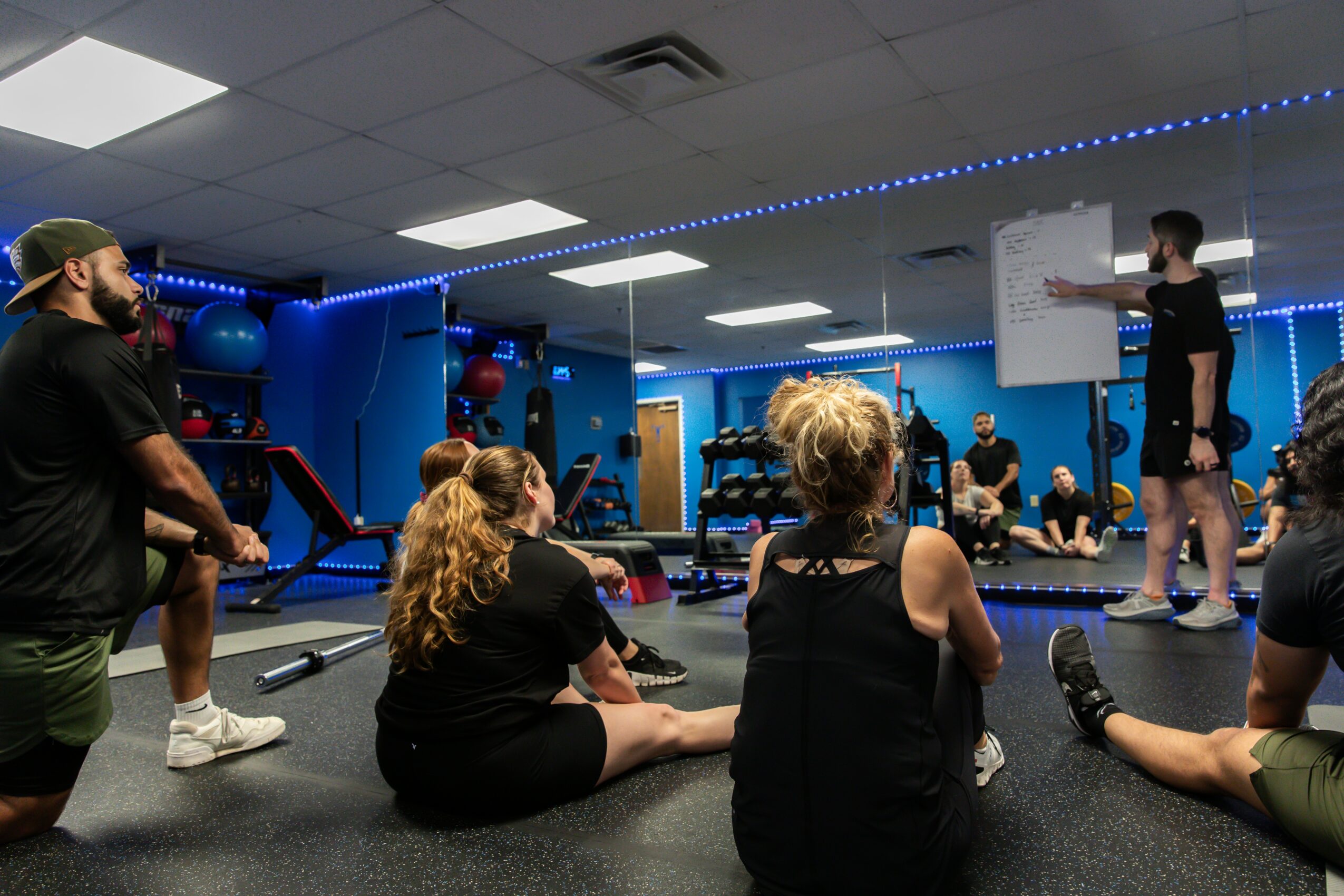 Group of people sitting on the floor listening to a person standing and presenting in a gym with blue walls and exercise equipment.