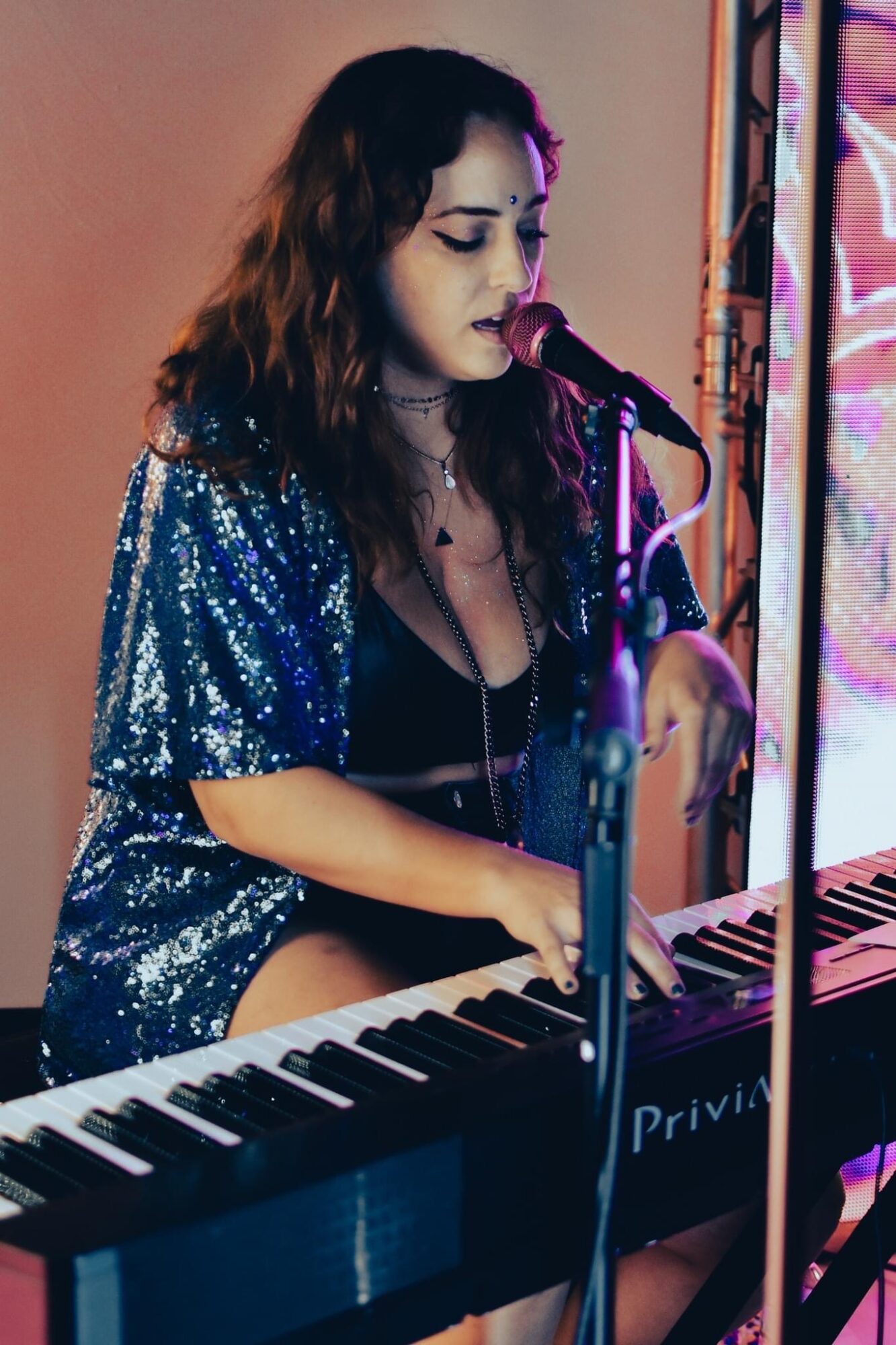 Woman with dark wavy hair singing into a microphone while playing a keyboard, wearing a shiny blue jacket.