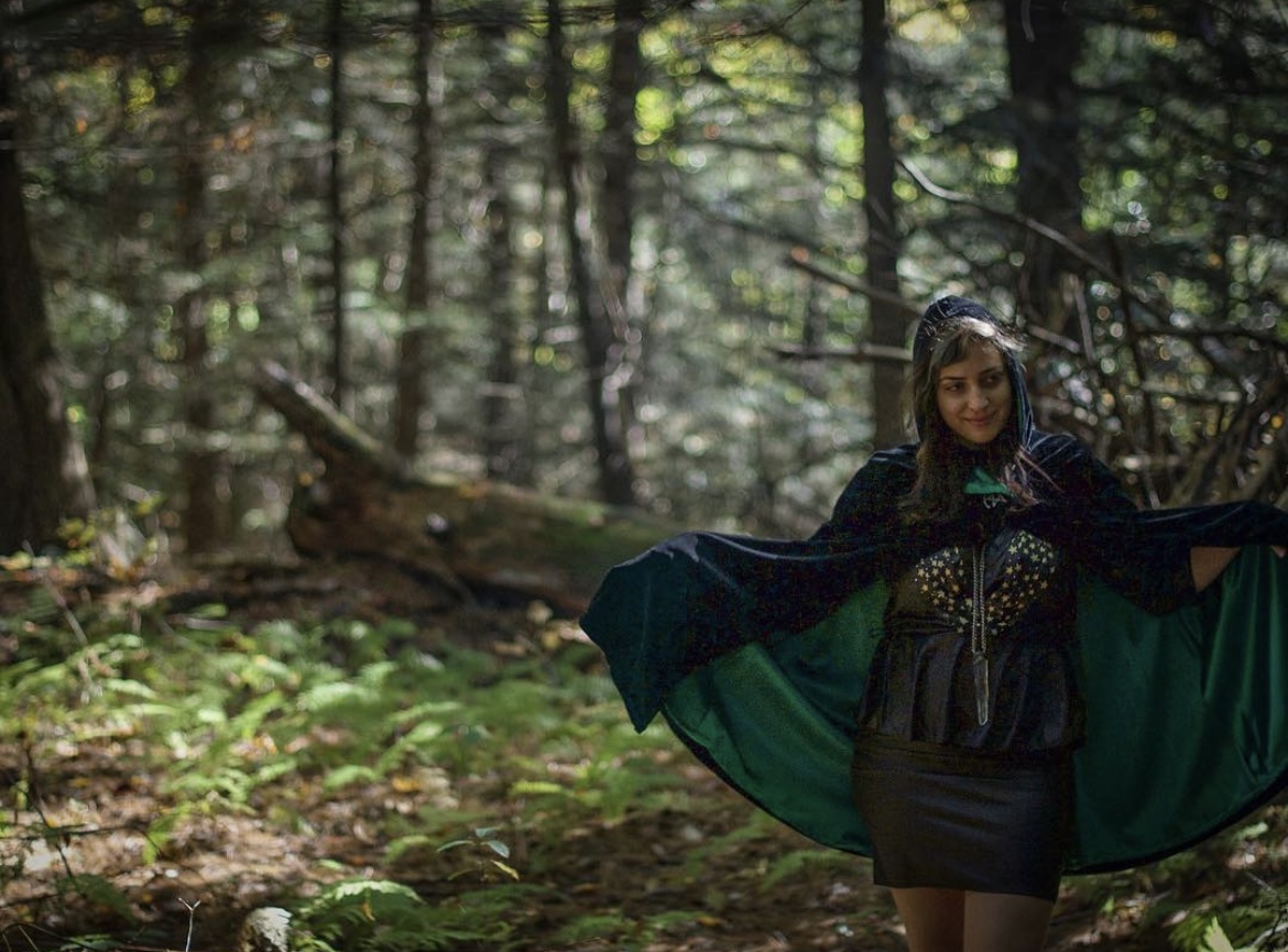 Woman in a forest with trees and greenery, wearing a dark outfit and holding a flowing green fabric.