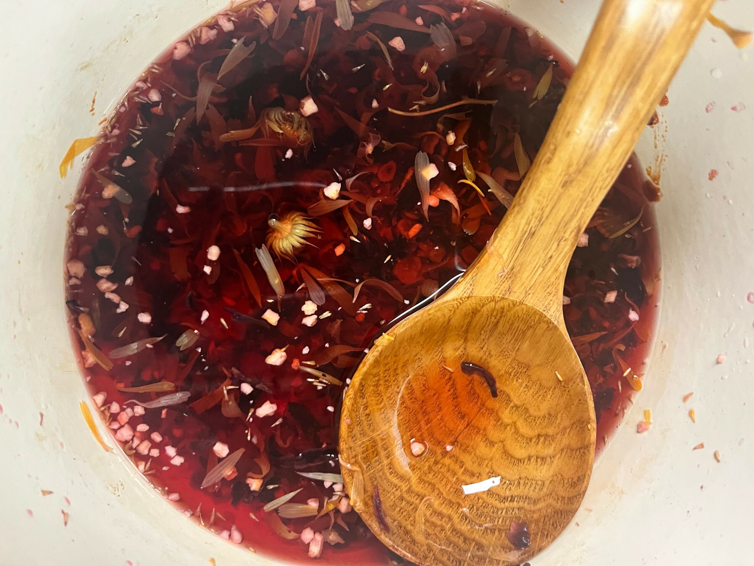A bowl of dark red liquid with floating ingredients and a wooden spoon inside, viewed from above.
