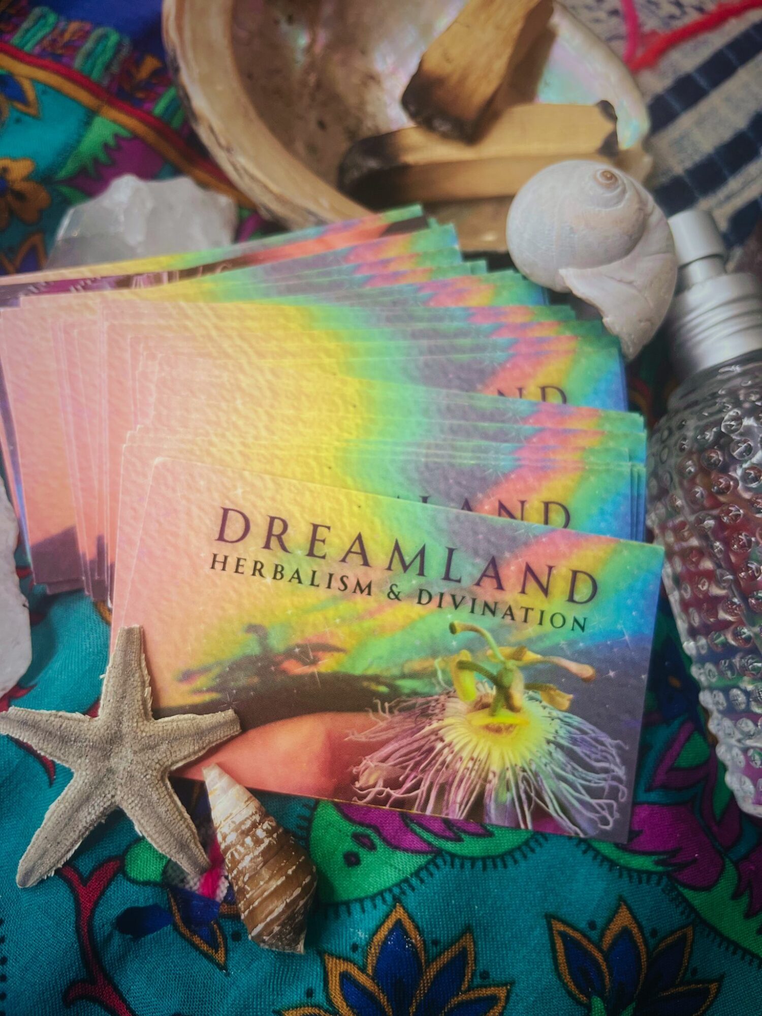 Colorful Dreamland herbalism and divination cards, starfish, seashells, and a water bottle on a vibrant fabric background.