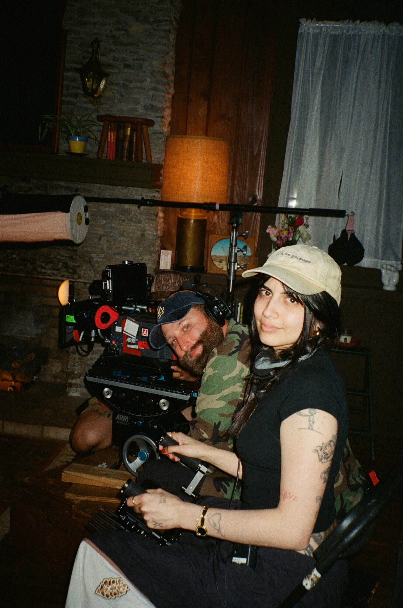 Two people, one with a cap and tattoos, sitting in a cozy room with warm lighting and a window with curtains.