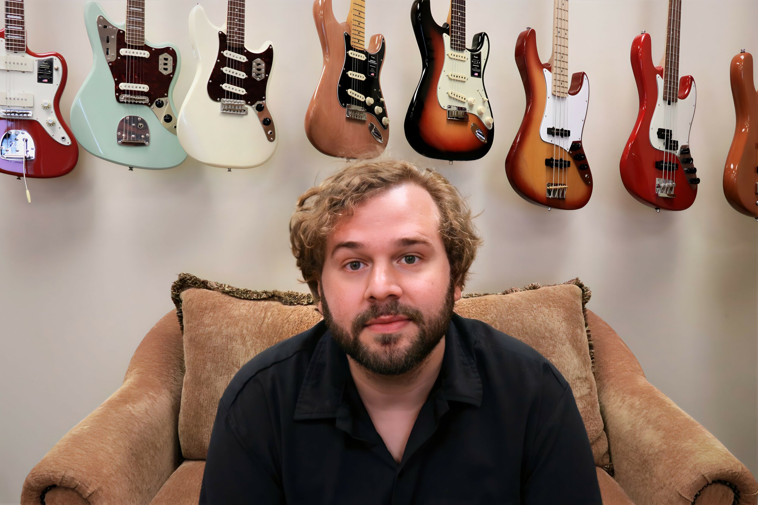 Man with curly hair and beard sitting on a sofa in front of a wall with electric guitars hanging.