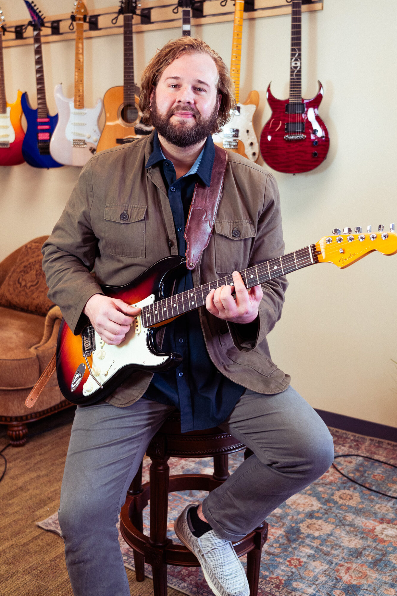 Man with beard and curly hair playing electric guitar in music store with guitars on wall behind him.