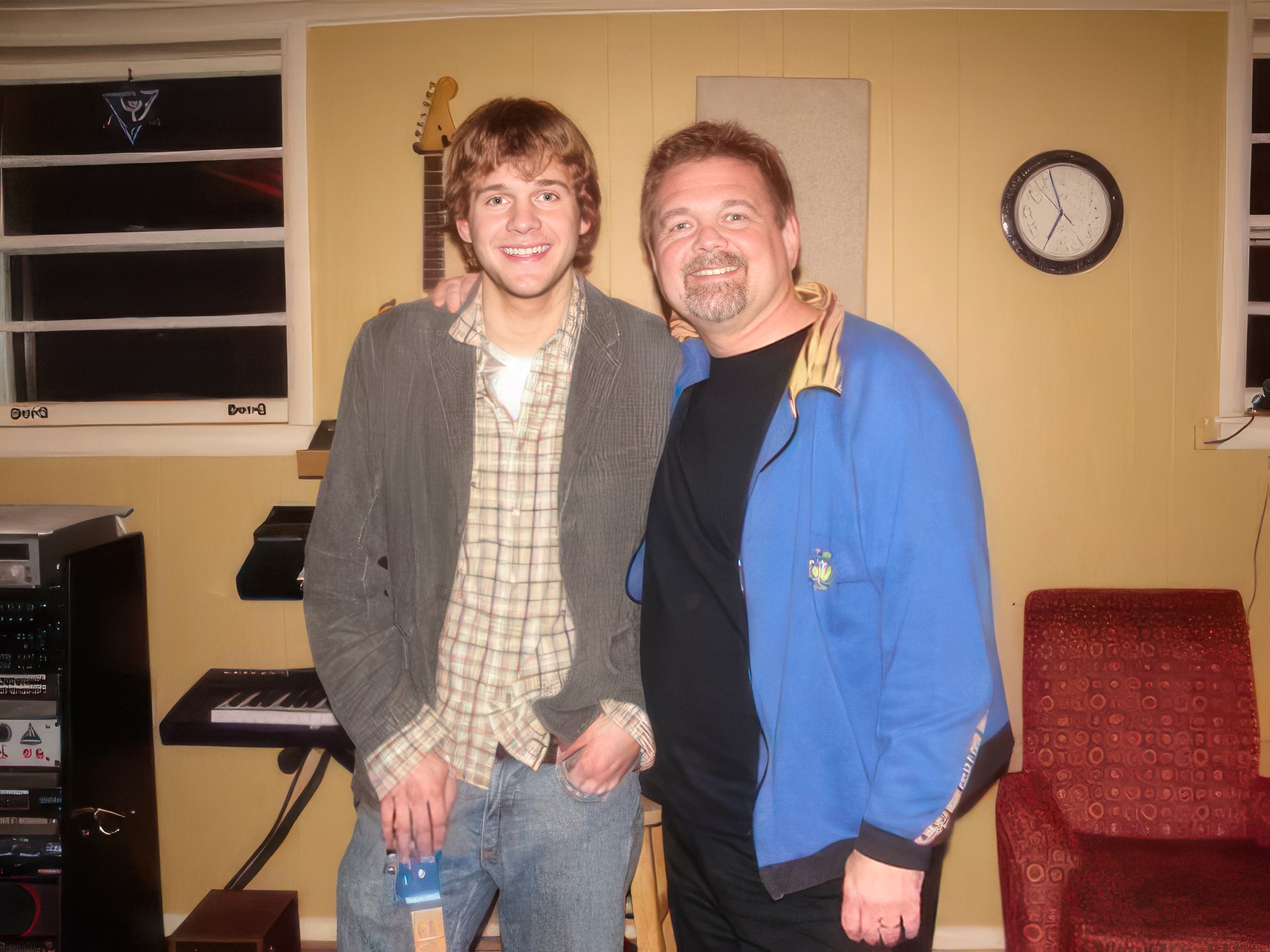Two men standing indoors, smiling, with musical instruments and a clock on the wall behind them.