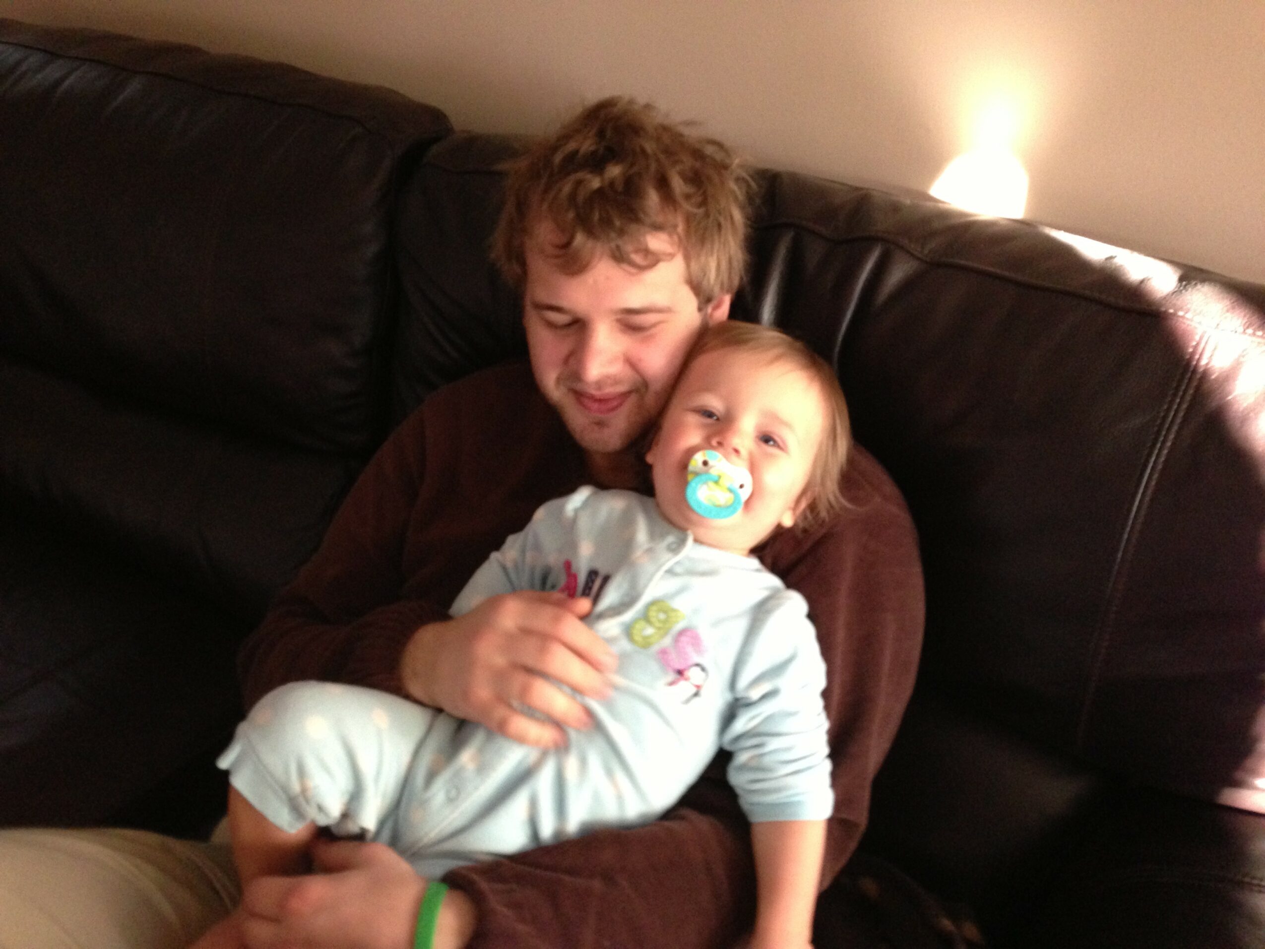Man with curly hair holding a baby with a pacifier, sitting on a dark sofa in a cozy room.