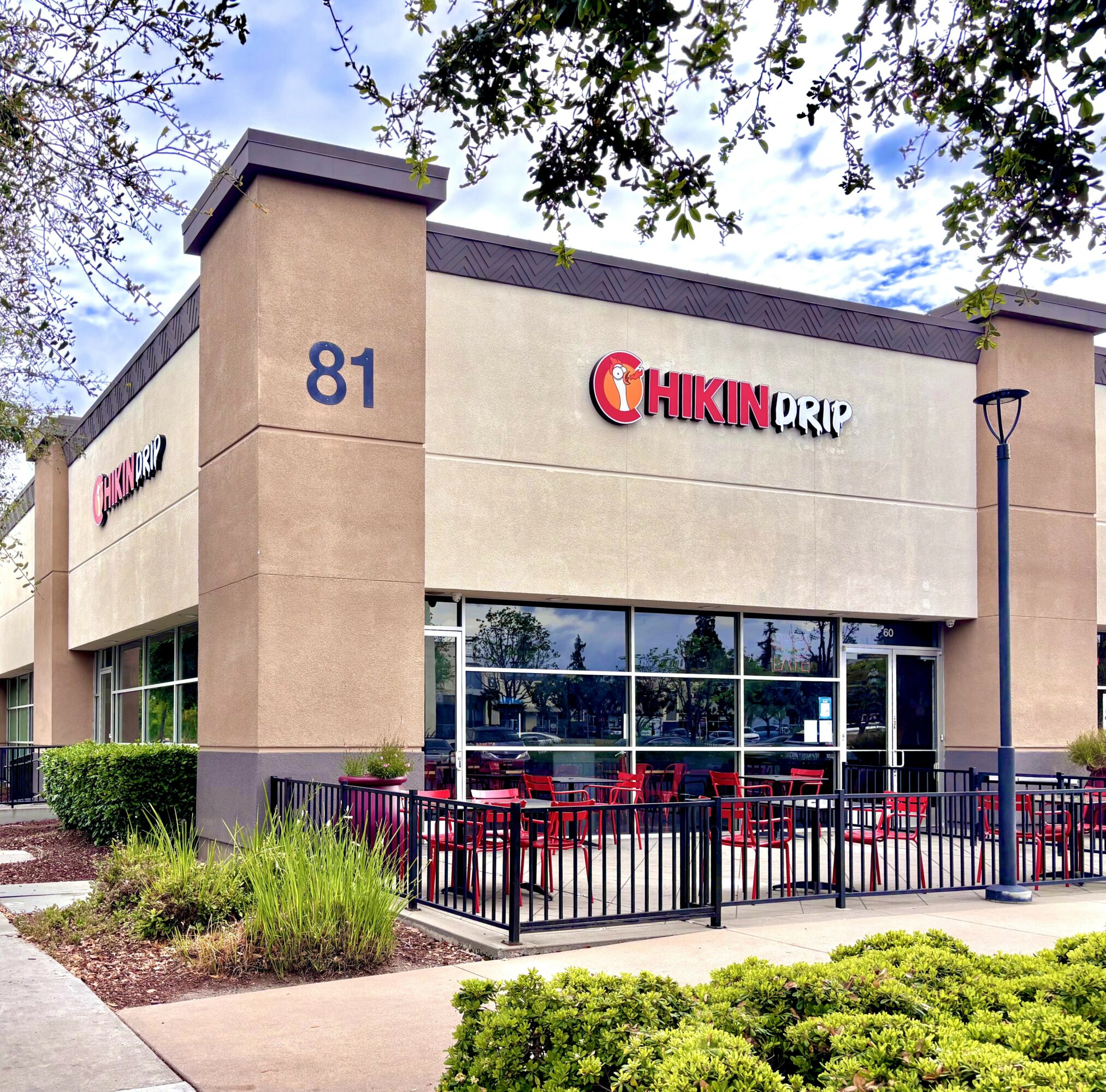 Chikin Drip restaurant building with large windows, outdoor seating area, and greenery, under a partly cloudy sky.