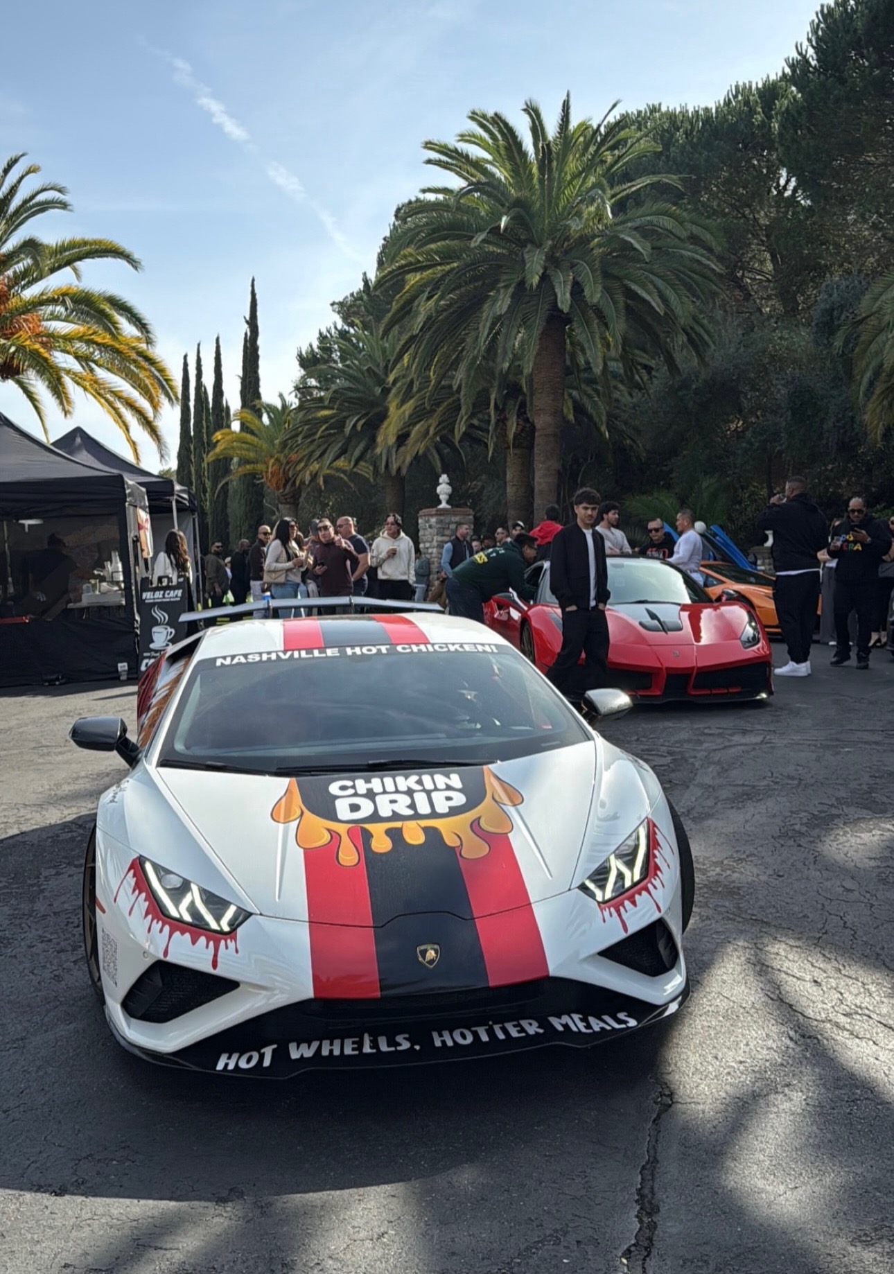 Silver sports car with black and red stripes, surrounded by people and other cars outdoors, palm trees in background.