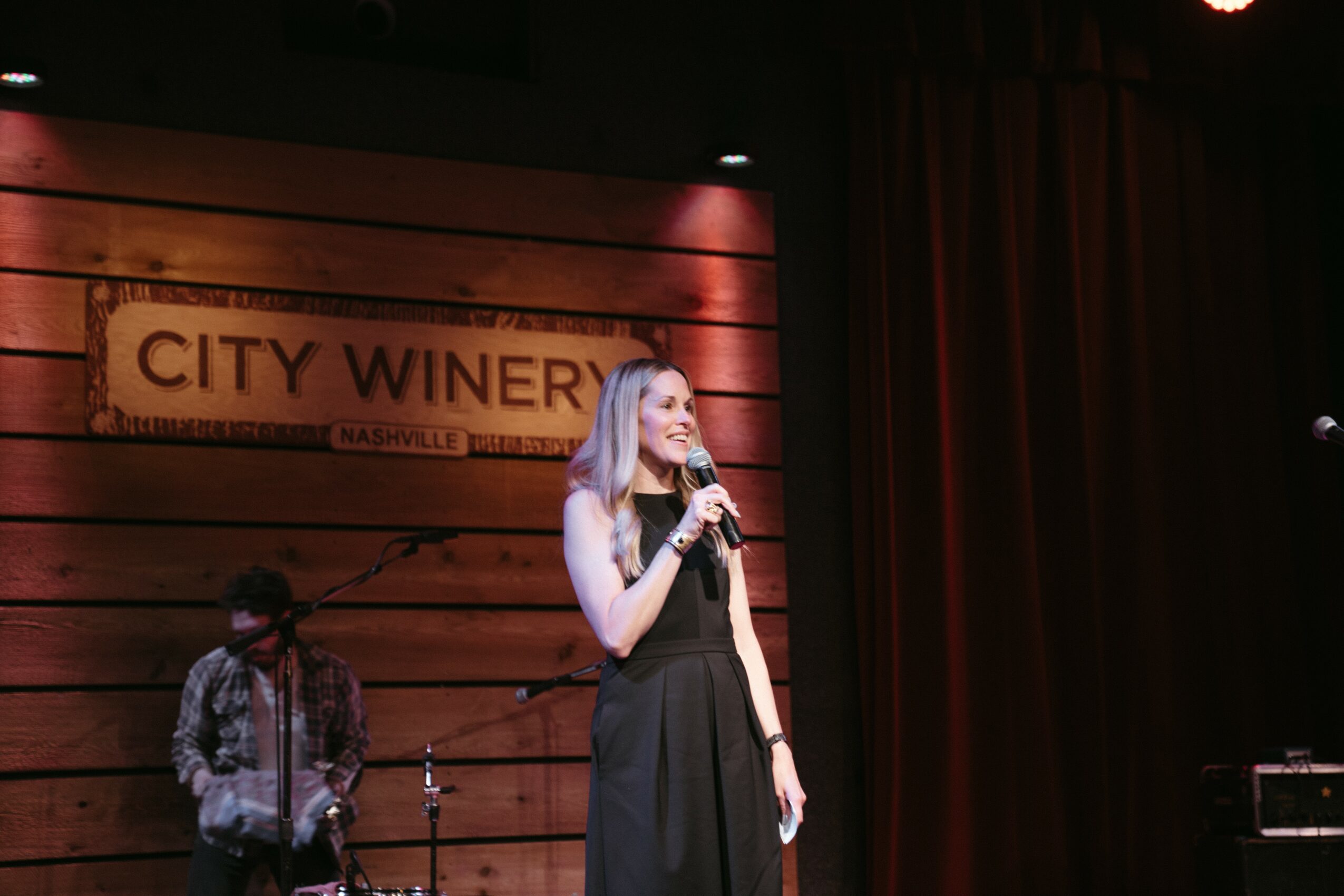 Woman with long hair holding a microphone on stage, with a man in the background, in front of a wooden wall with a sign.