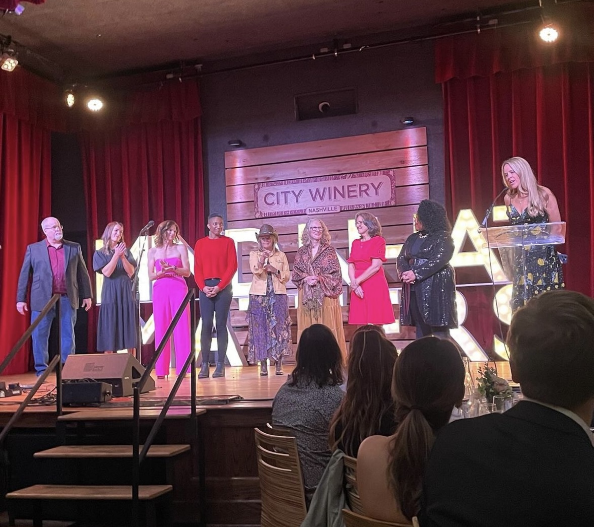 Group of people on stage at City Winery, some holding microphones, audience seated watching, red curtains in background.