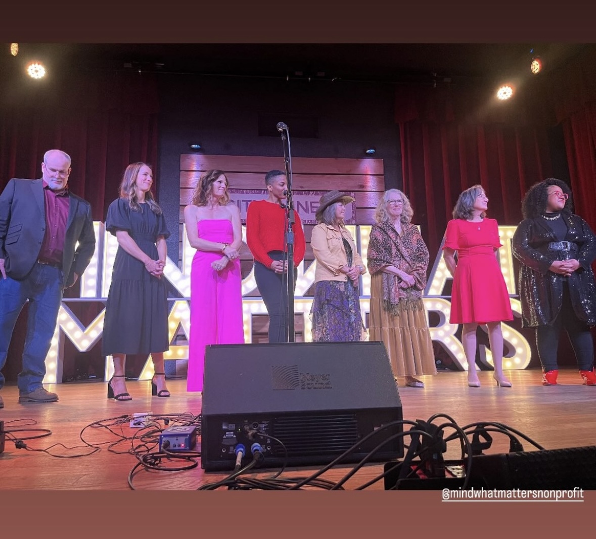 Group of eight people standing on stage with red curtains, stage lights, and a microphone stand behind them.