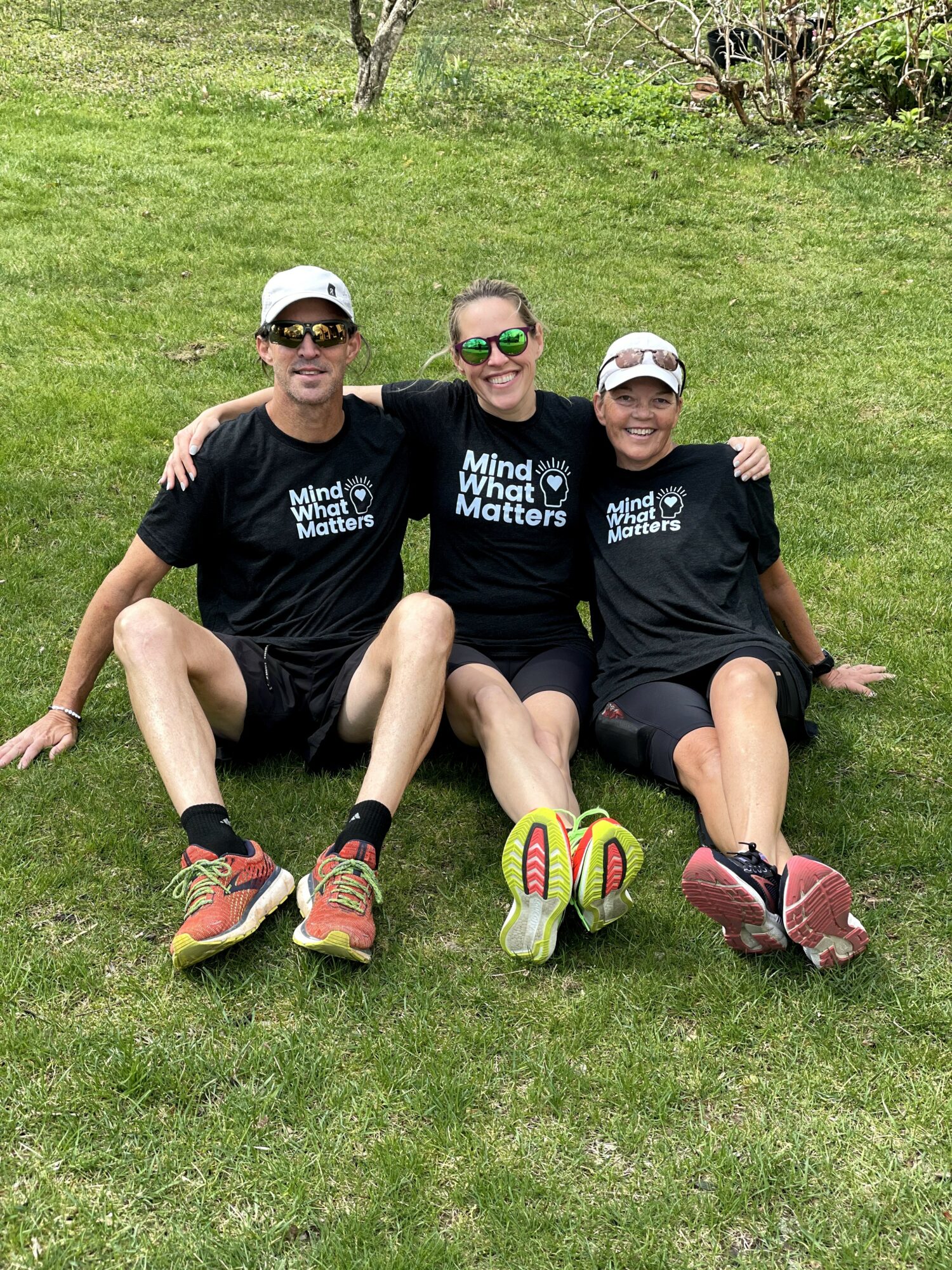 Three people sitting on grass, smiling, wearing black shirts with white text, and sunglasses, with trees in background.