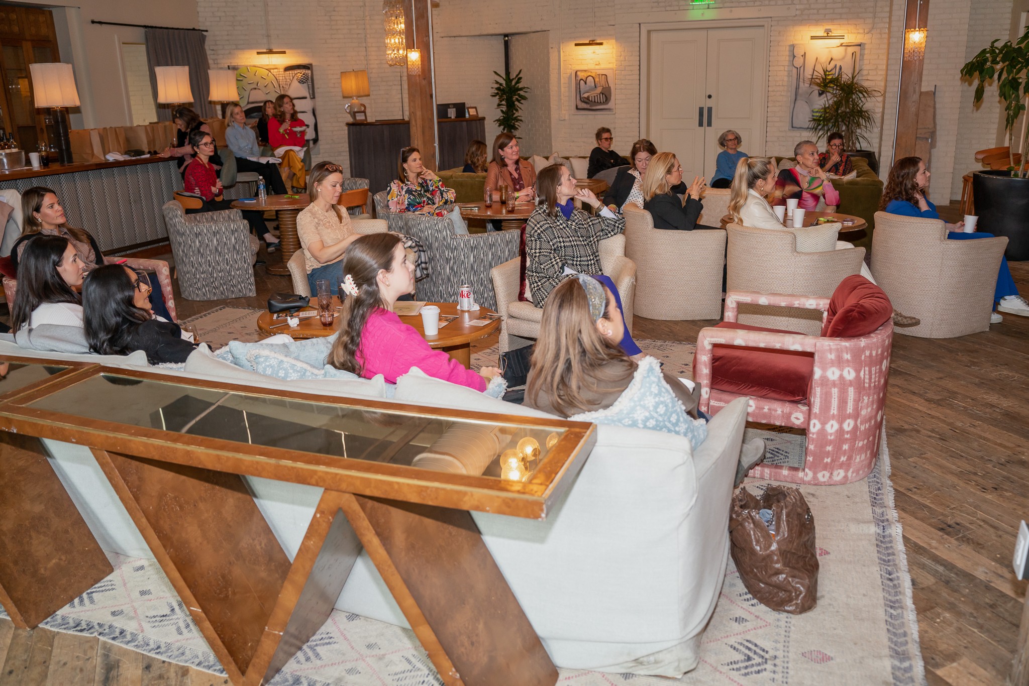 People seated at tables and couches in a spacious, well-lit room with wooden floors and decorative plants.
