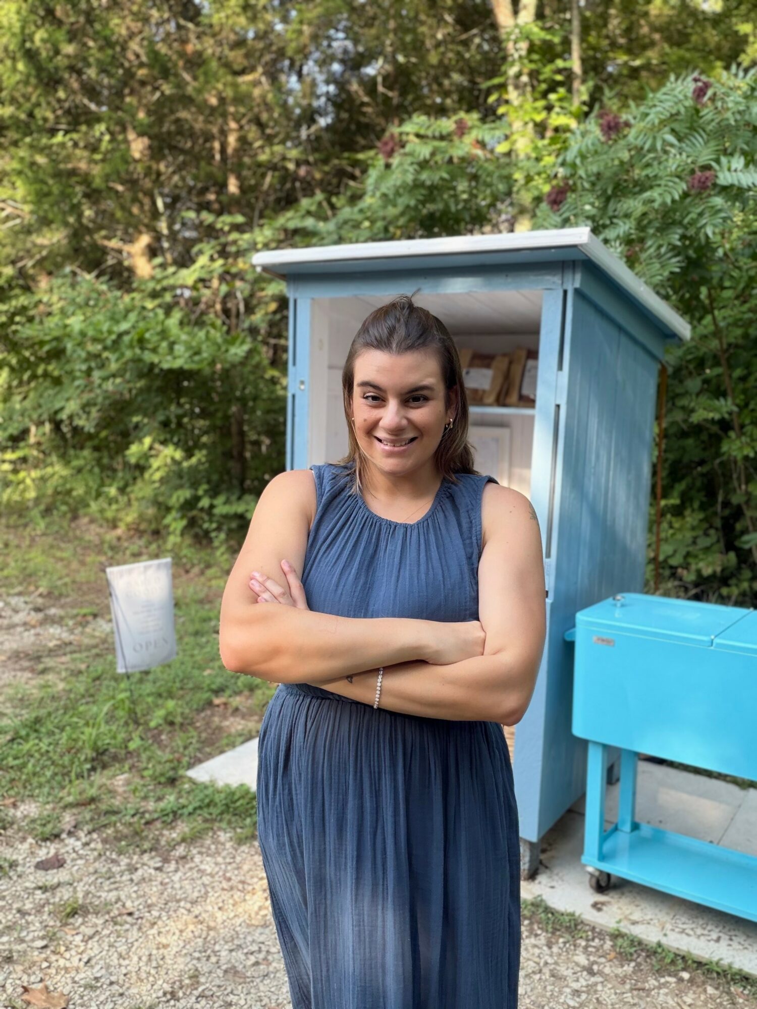 Smiling woman in a blue dress standing outdoors with arms crossed, behind a blue wooden structure and greenery.