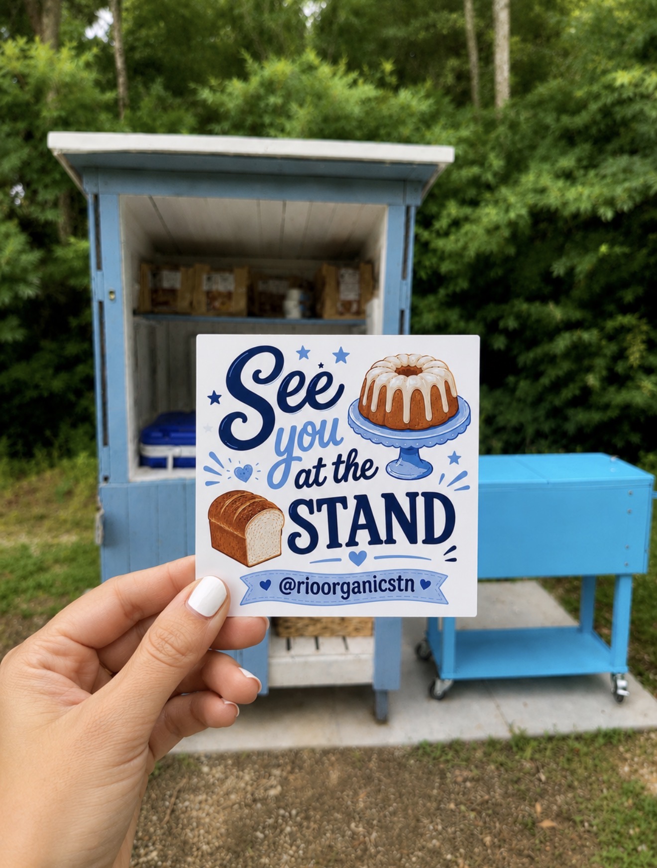 Person holding a sign with cake and chocolate, reading 'See you at the stand,' in front of a blue outdoor stand, outdoors.