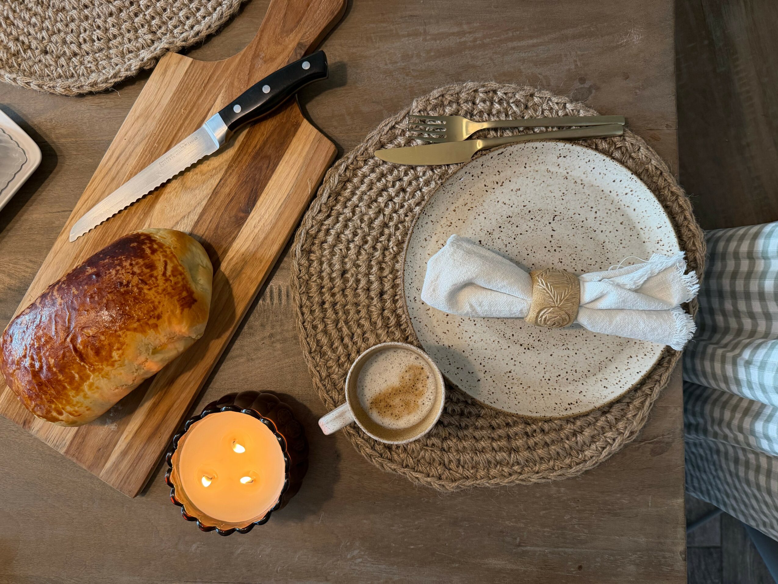 Table setting with bread, knife, cutting board, cup, and plates on woven placemats, viewed from above.