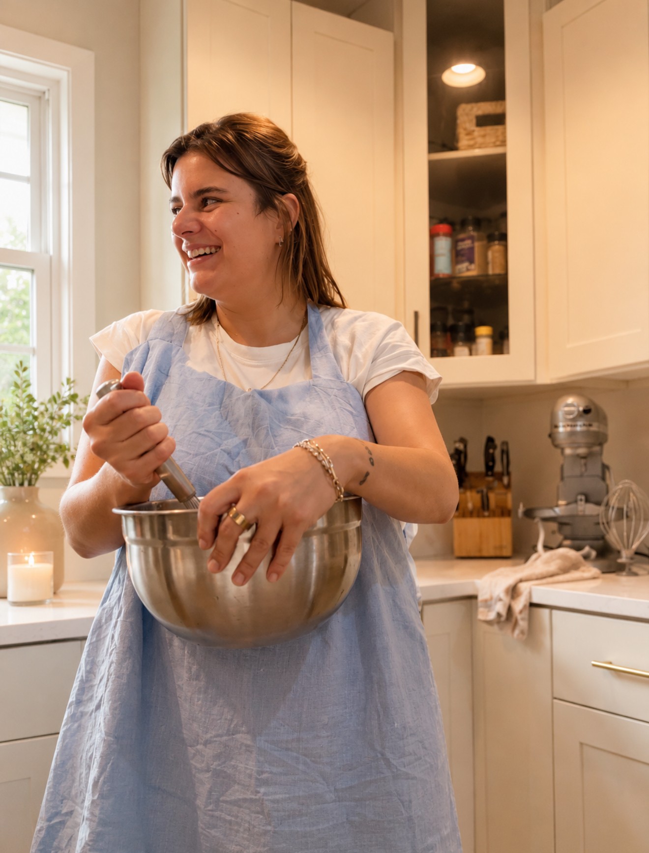 Woman smiling in kitchen, holding a mixing bowl, wearing a white shirt and blue apron, with cabinets and kitchen items in background.