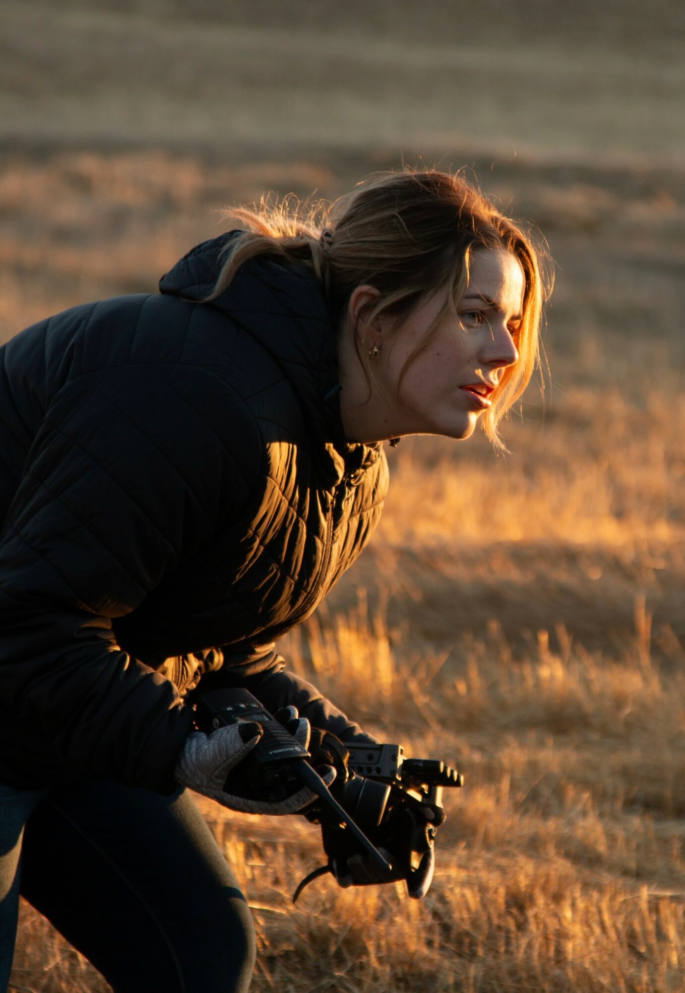 Woman in black jacket holding a camera, outdoors in a field during sunset, looking to the side.