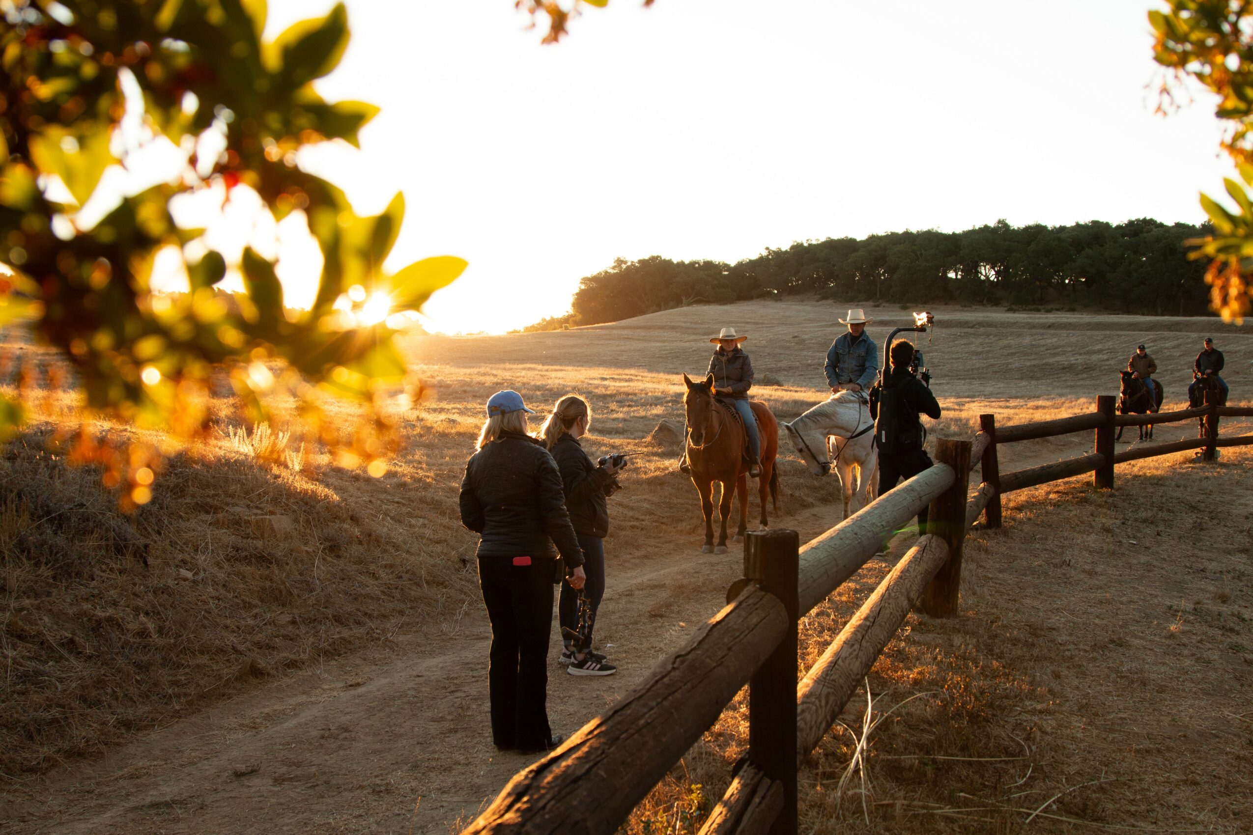People walking and riding horses along a dirt path with a sunset in the background, trees on the side, and a wooden fence.