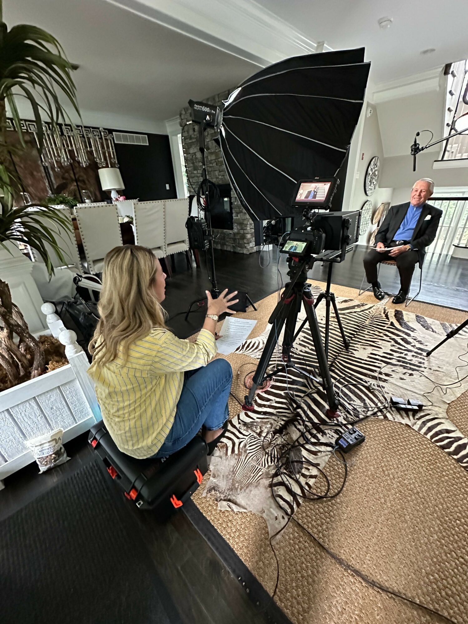 Person in yellow sweater operating camera equipment in a room with zebra print rug and plants.