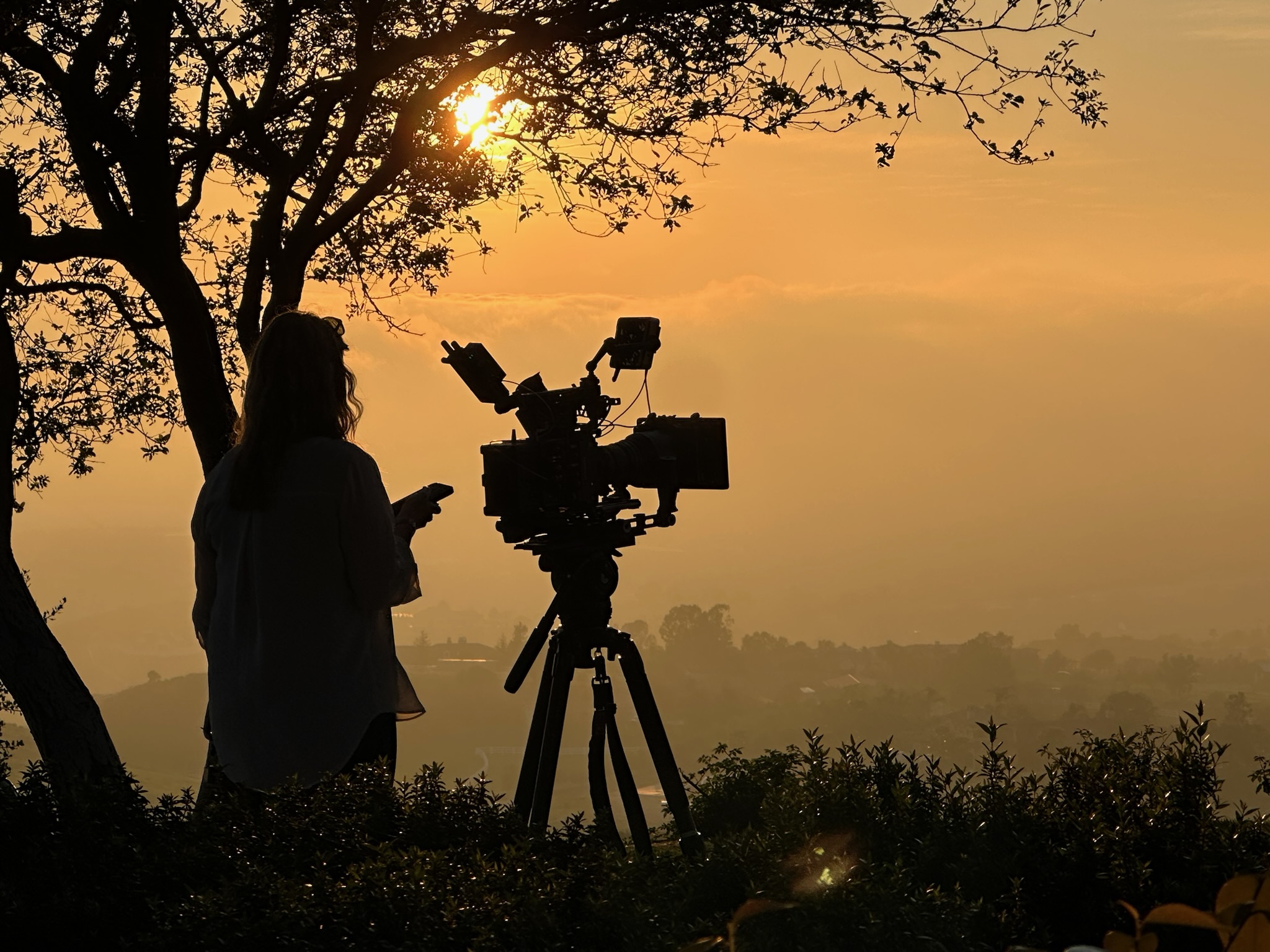 Person standing outdoors with camera on tripod during sunset, holding a device, silhouette against sky.