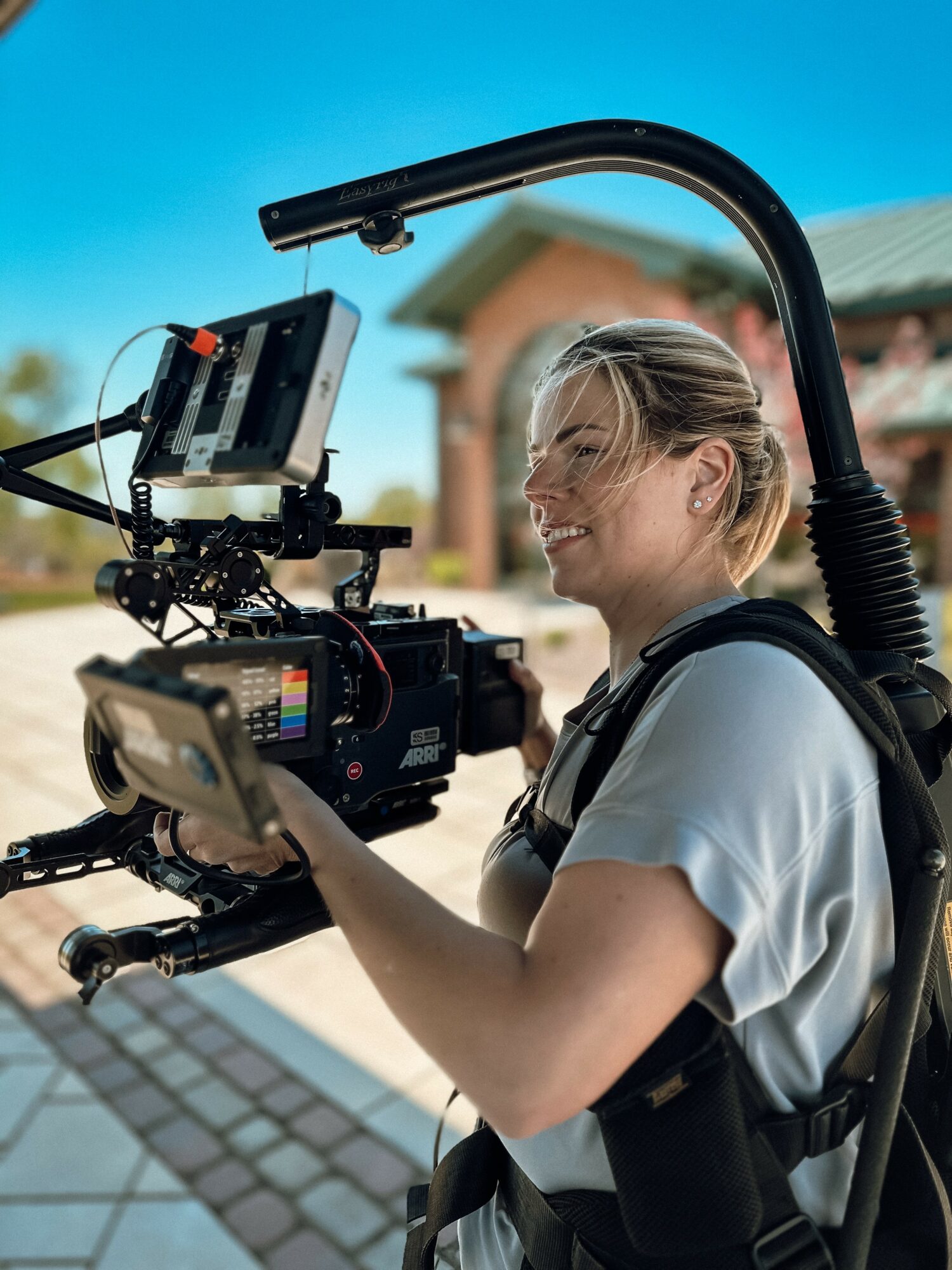 Woman operating professional camera equipment outdoors with a building and blue sky in background.